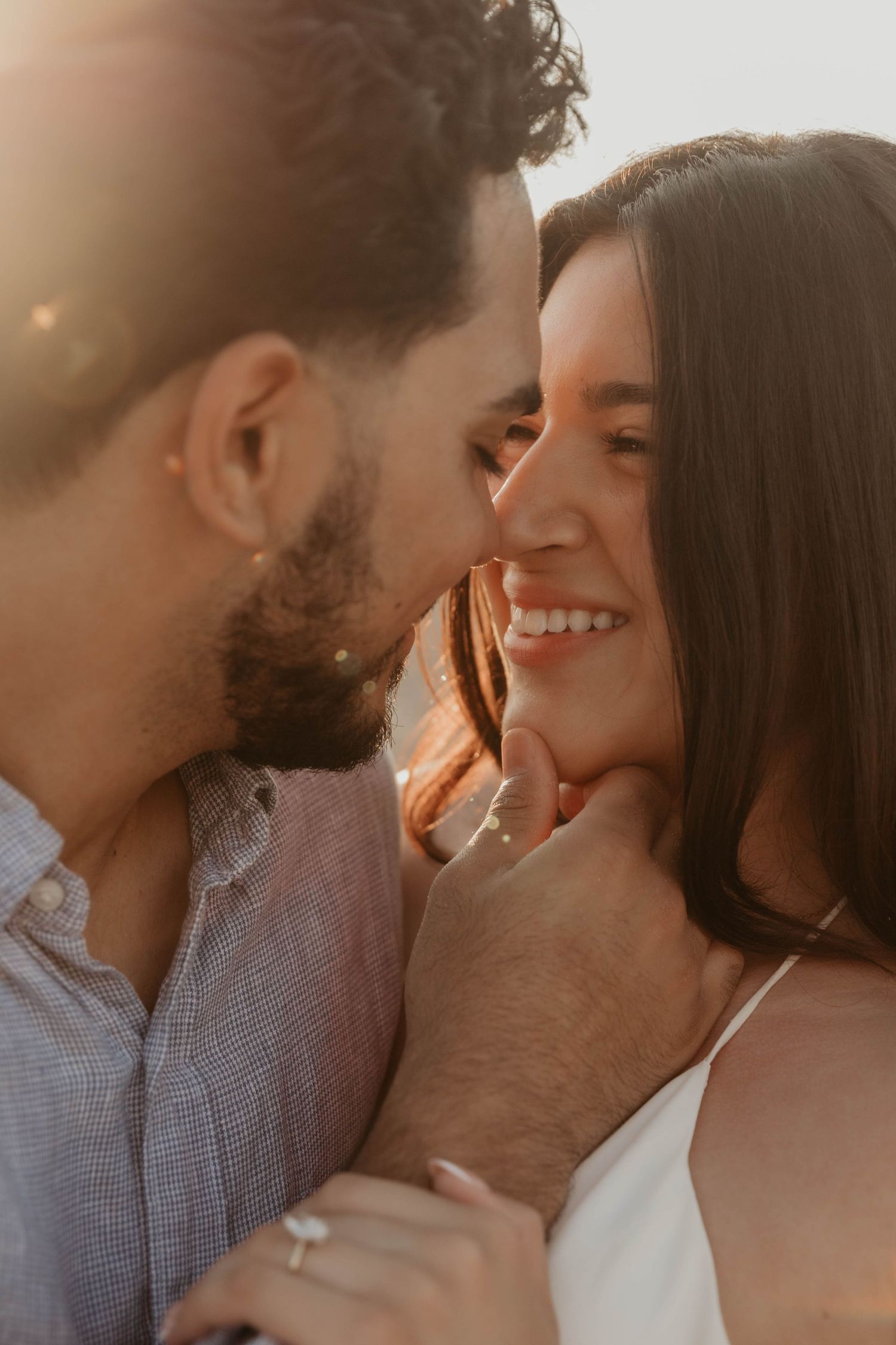 A couple shares a romantic golden hour moment, man gently holds woman's face as she smiles warmly wearing an engagement ring.