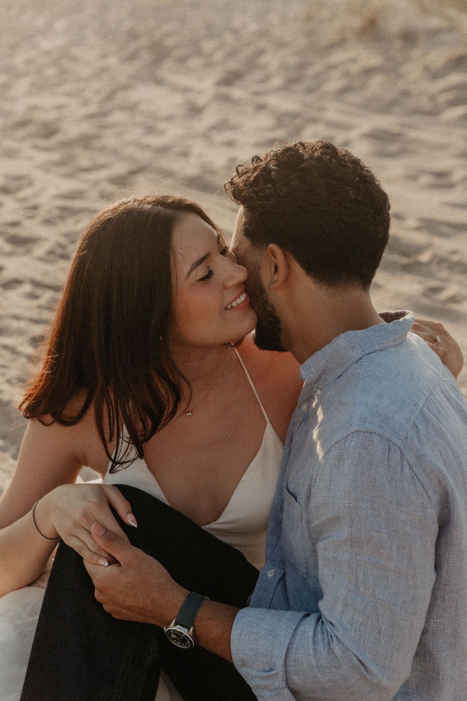 Couple sharing a romantic moment on a sandy beach at golden hour, woman smiling as man leans in close.