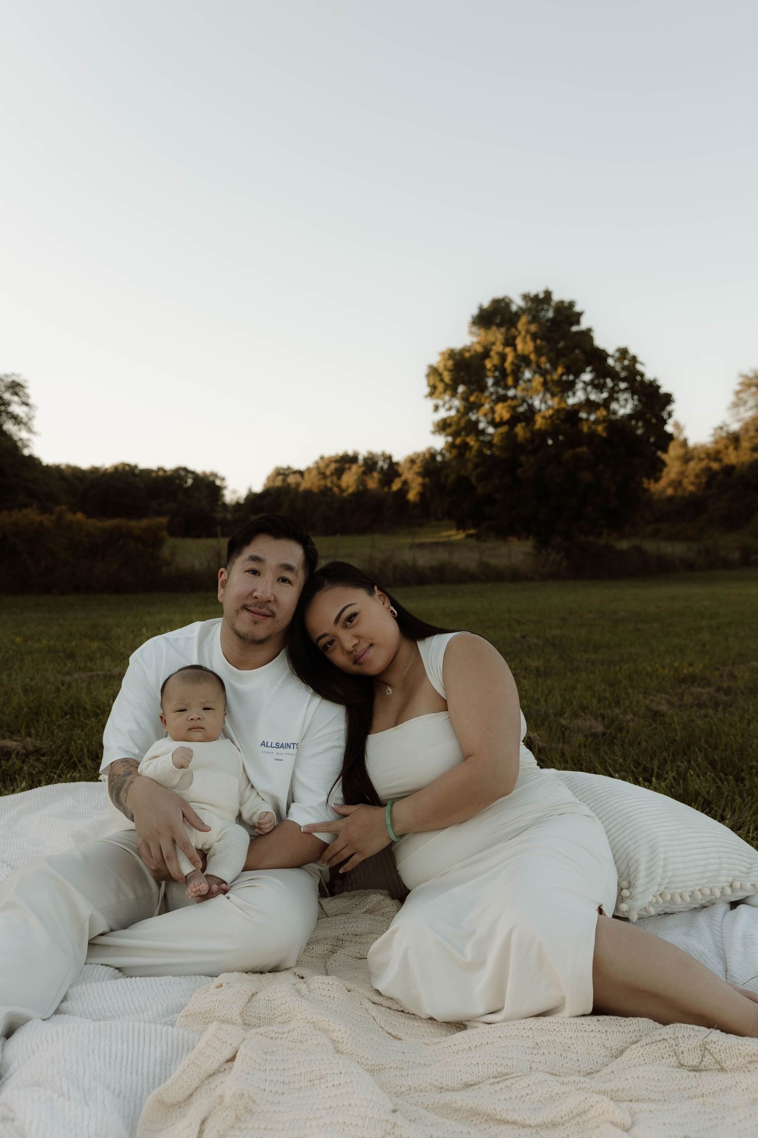A young family poses on a white blanket in a green field at golden hour, parents in white outfits cradling their newborn baby.