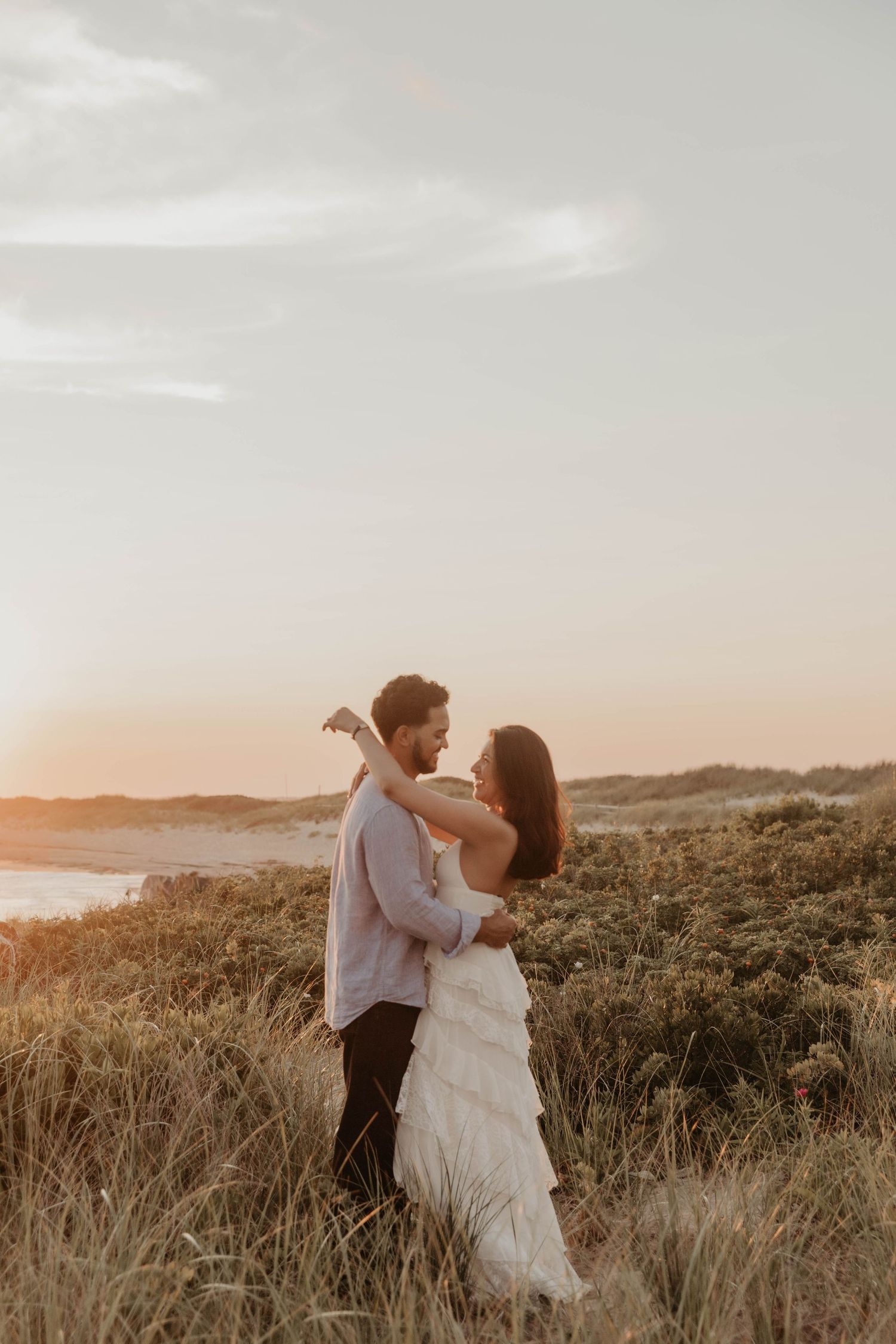 Couple embracing in golden sunset light among coastal dunes, bride in white ruffled gown and groom in casual attire.