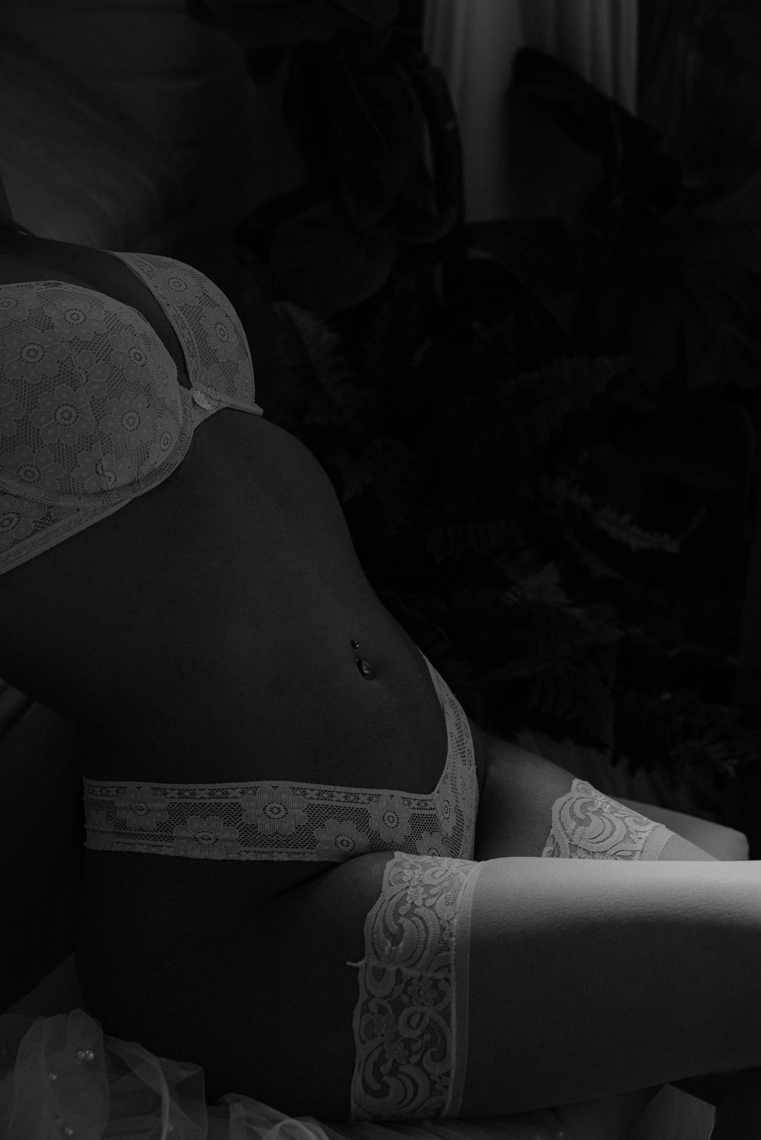 Black and white artistic close-up of a woman in lace lingerie and stockings among dark foliage.