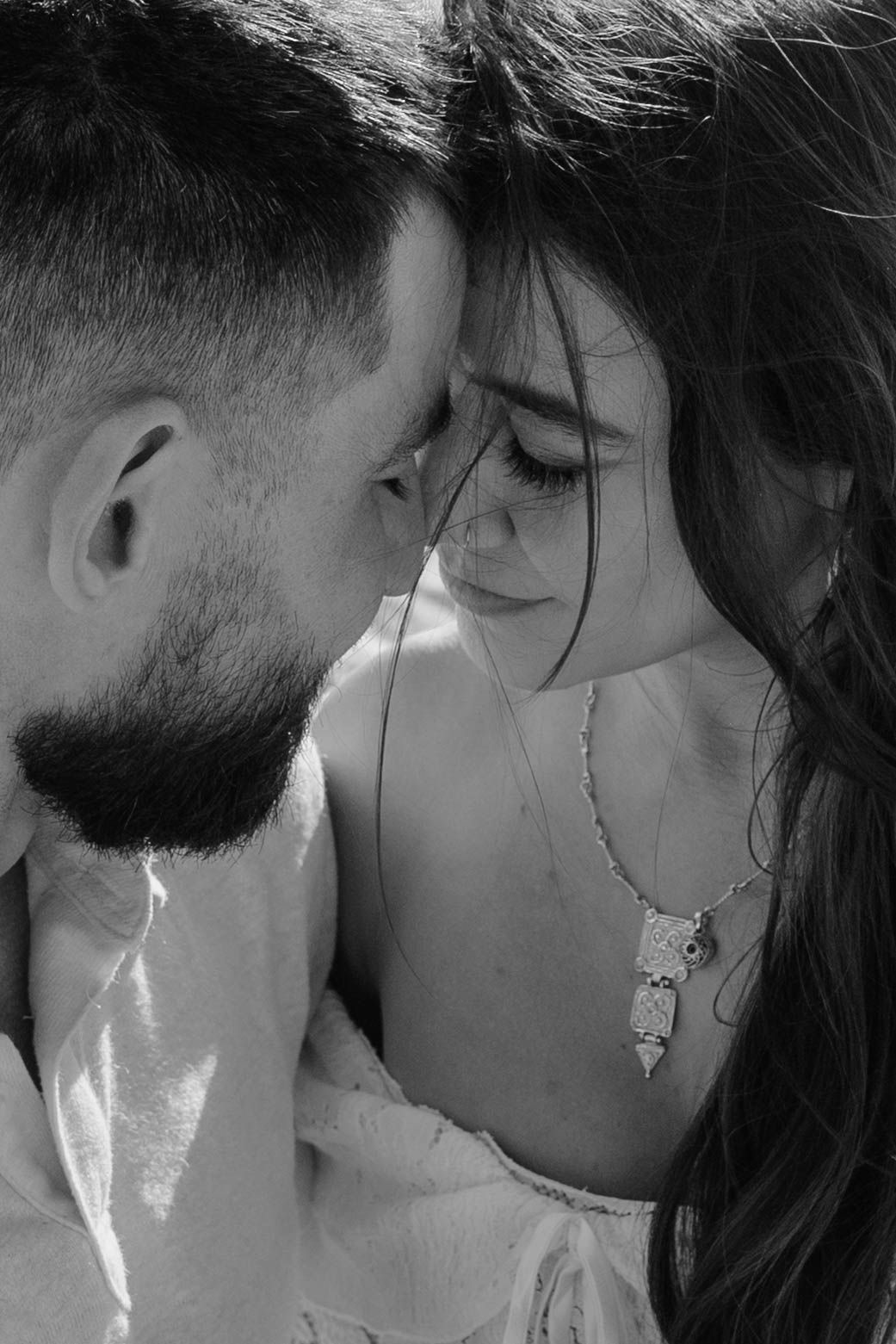 Black and white portrait of a bearded man and brunette woman with necklace sharing an intimate forehead-to-forehead moment.