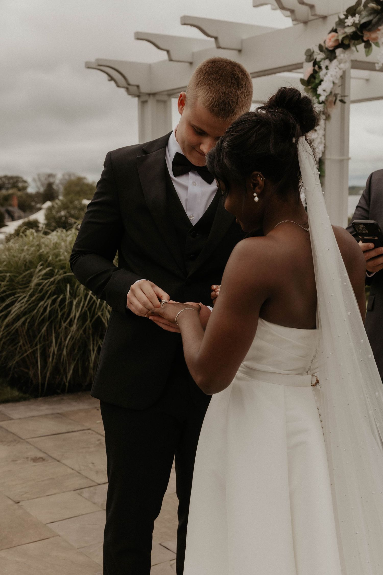 A groom in a black tuxedo and bow tie places a ring on his bride's finger during an outdoor wedding ceremony beneath a white pergola.