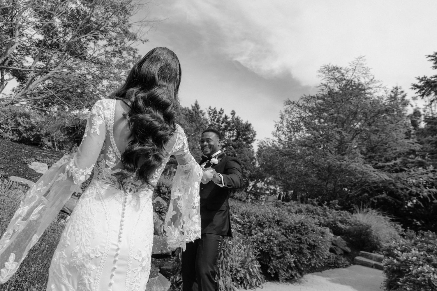 Black and white photo of bride in lace gown walking toward emotional groom in a lush garden setting.