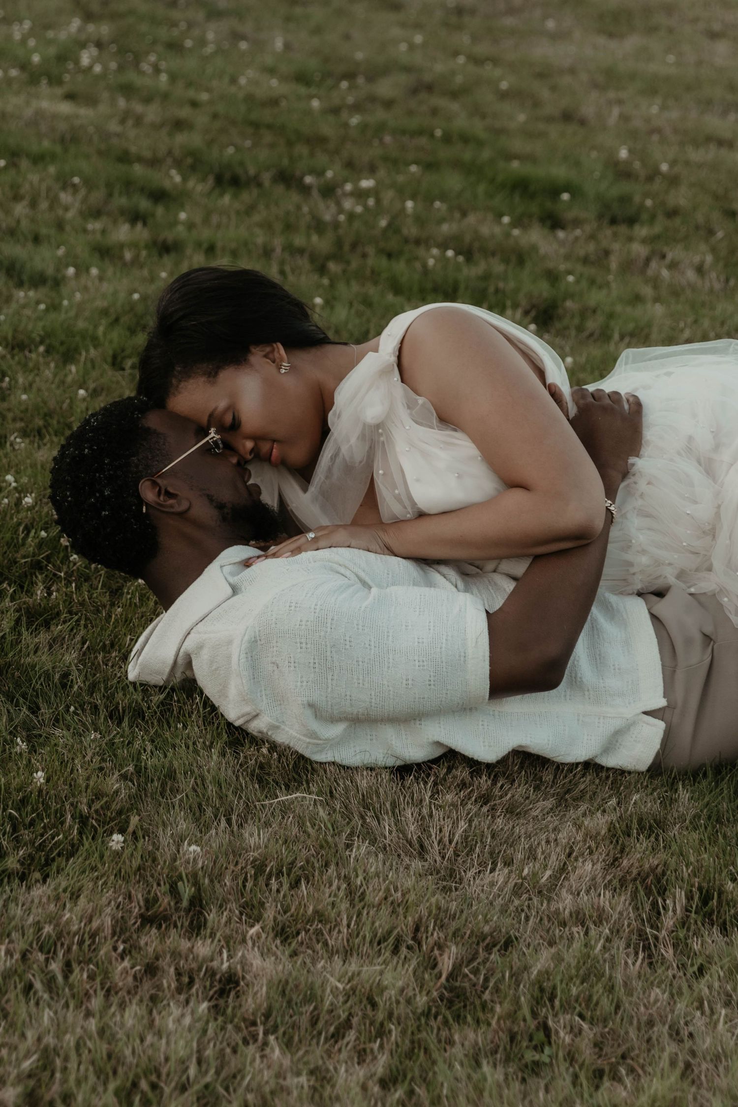 A couple sharing a romantic moment on green grass, woman in white dress leaning over man in light ivory shirt.