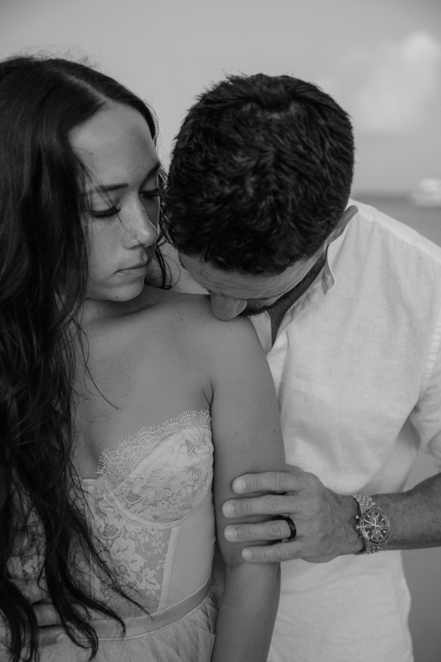 Black and white photo of a couple in an intimate embrace, man kissing woman's shoulder near the beach.