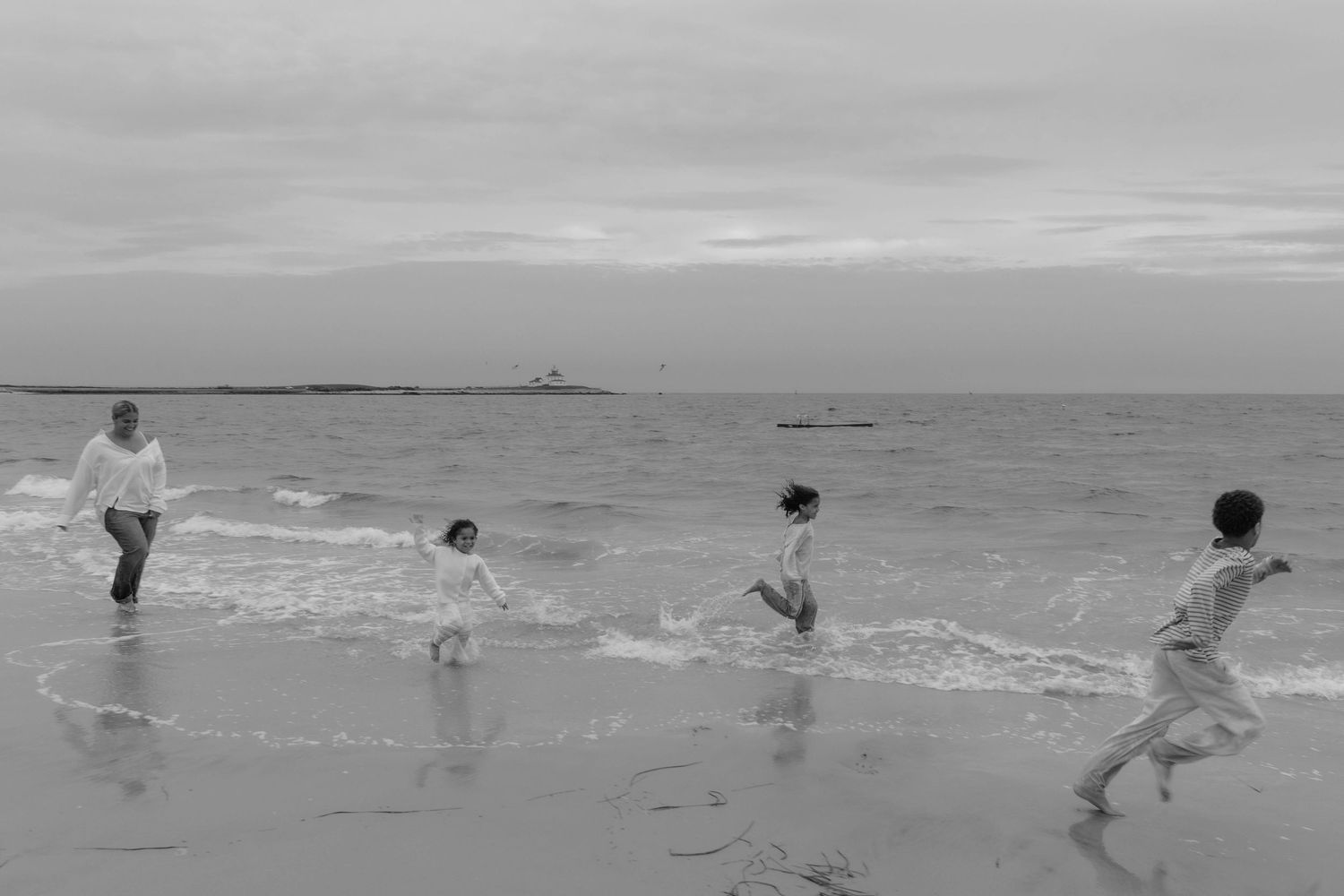 Black and white photo of family playing in ocean waves on a beach with a lighthouse visible in the distance.