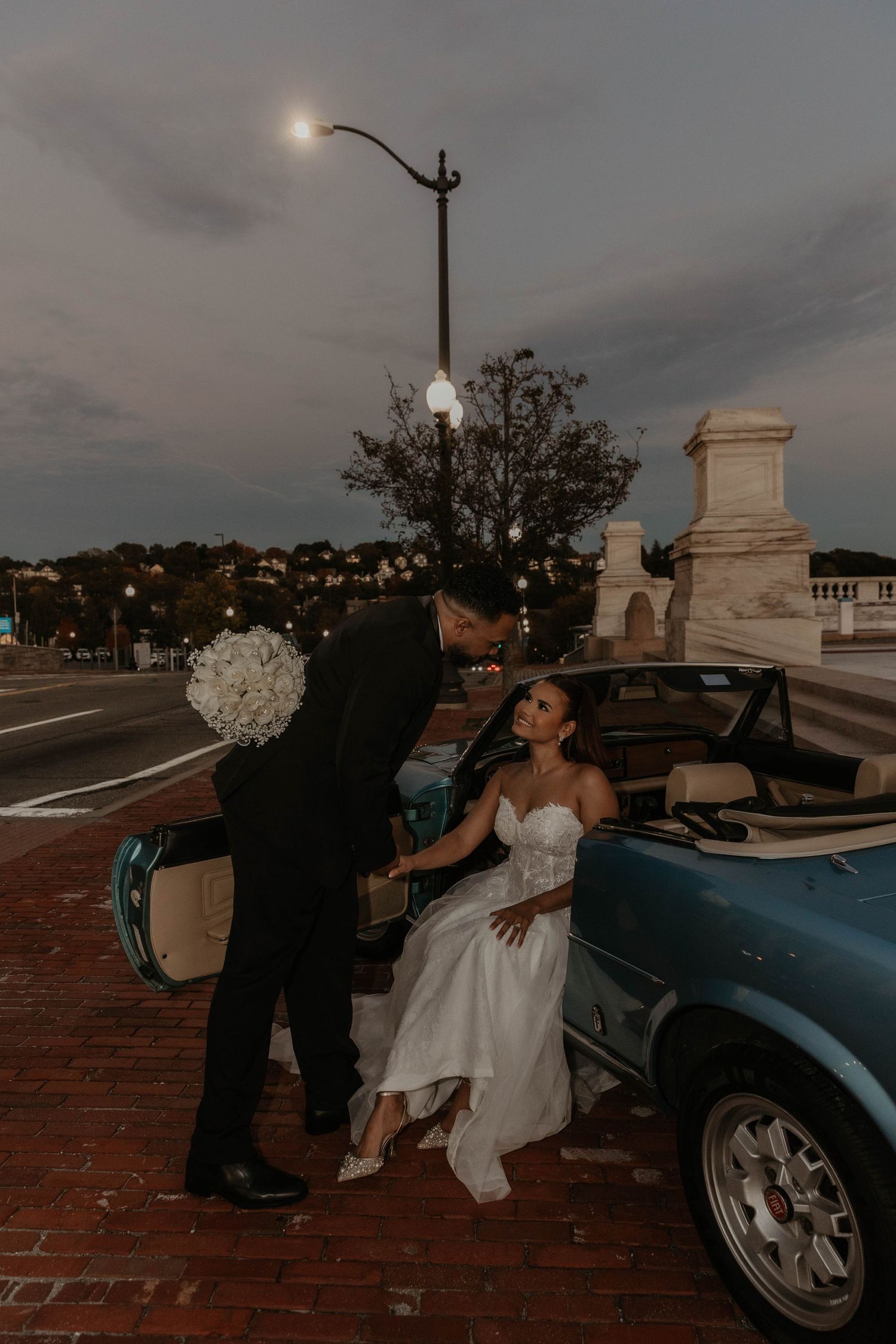 Bride in a white strapless gown sits in a vintage blue convertible as the groom leans in holding a bouquet at dusk beneath a street lamp.