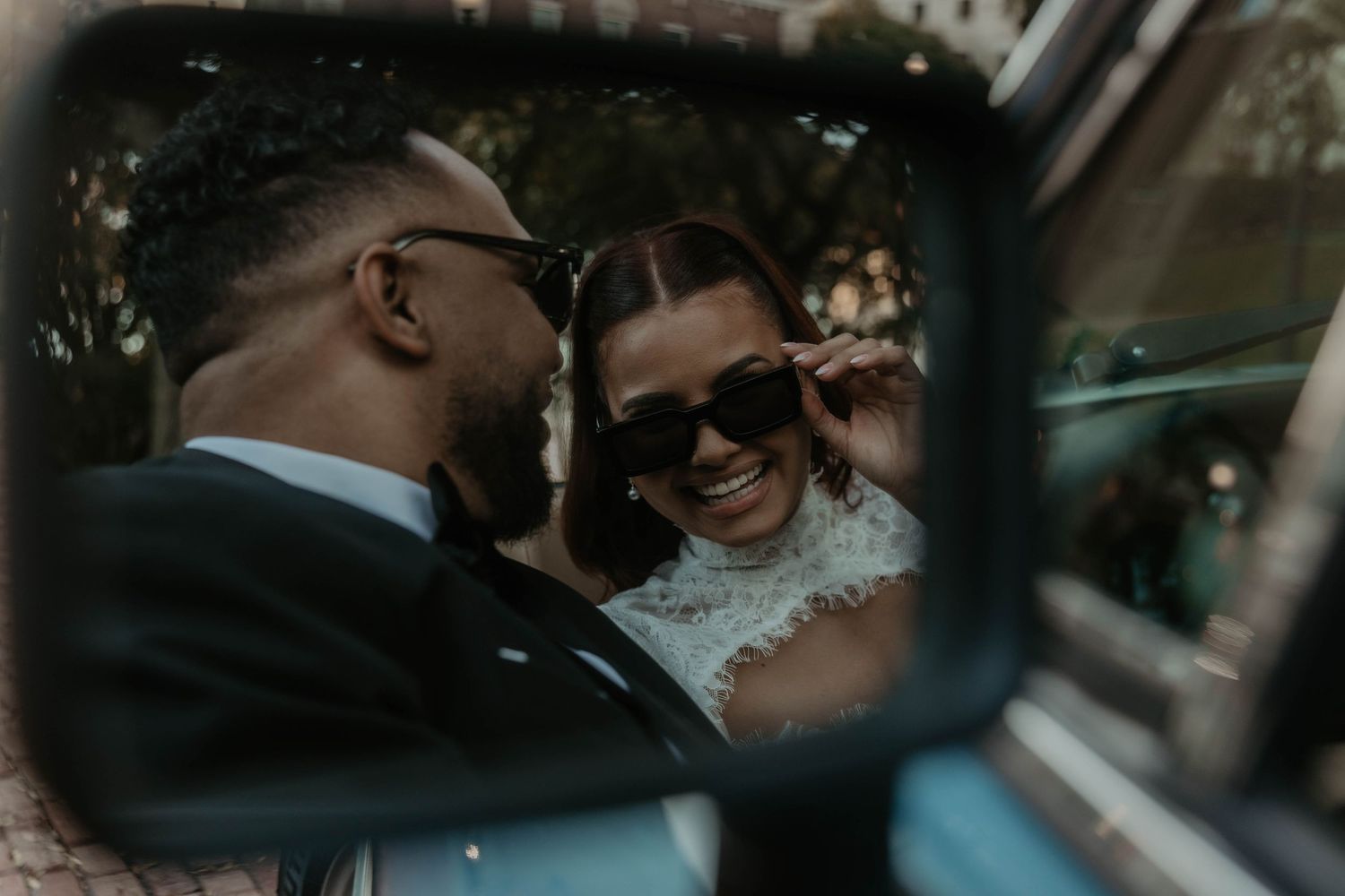 Reflection in a vintage car side mirror of a smiling woman lowering her sunglasses as a man in a dark suit leans in close.