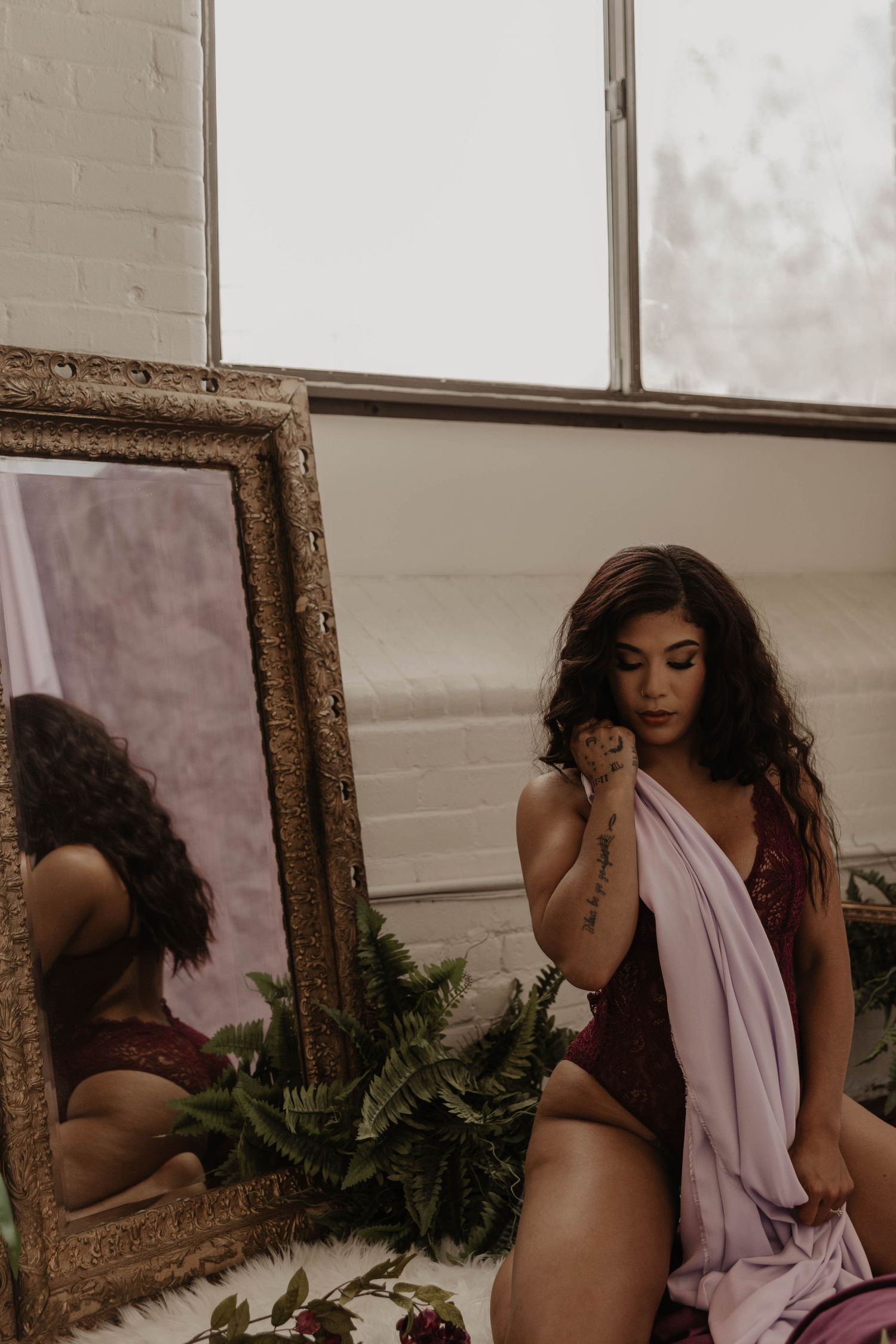 Woman in a burgundy lace bodysuit sits draped in a lavender wrap beside an ornate gold mirror, ferns and a bright window behind her.