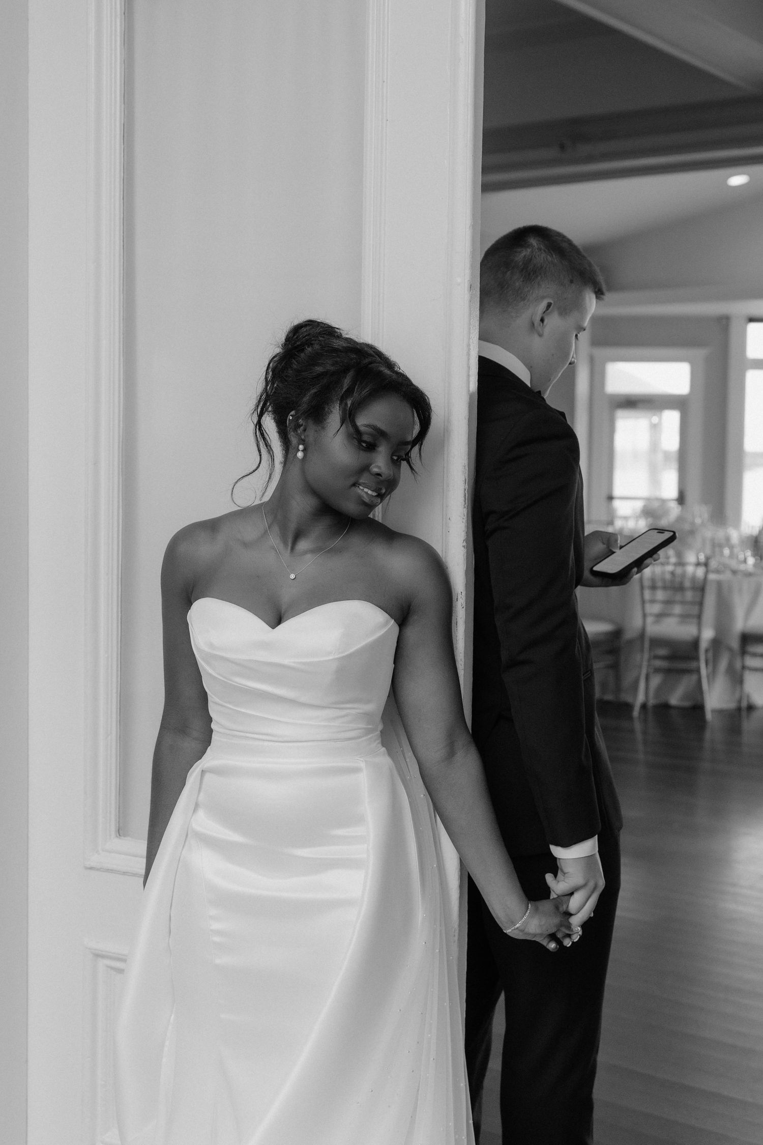 Black and white wedding photo of bride in strapless gown holding grooms hand around a doorframe.