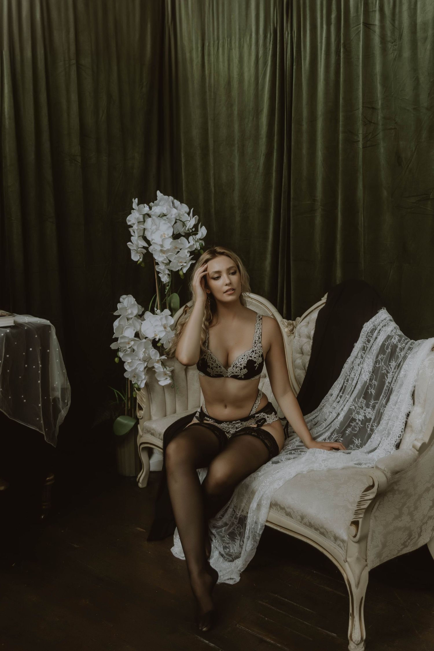 Woman in black lace lingerie sitting on ornate chair beside white orchids against green velvet curtains.