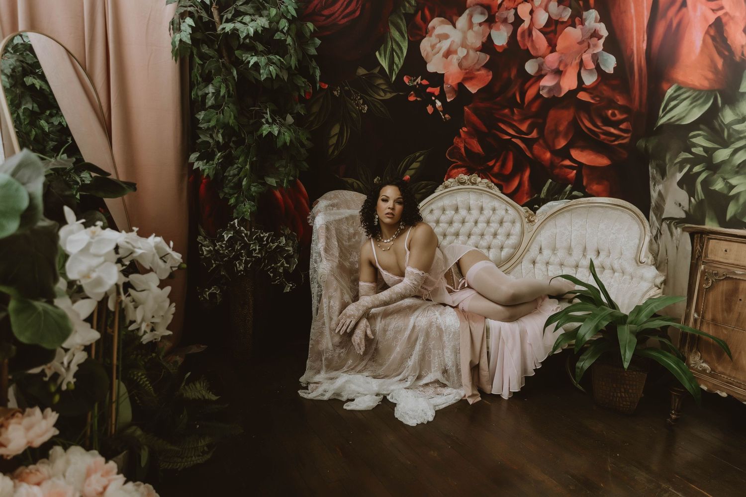 Woman in blush gown reclines on white vintage sofa surrounded by lush florals and dramatic floral backdrop.