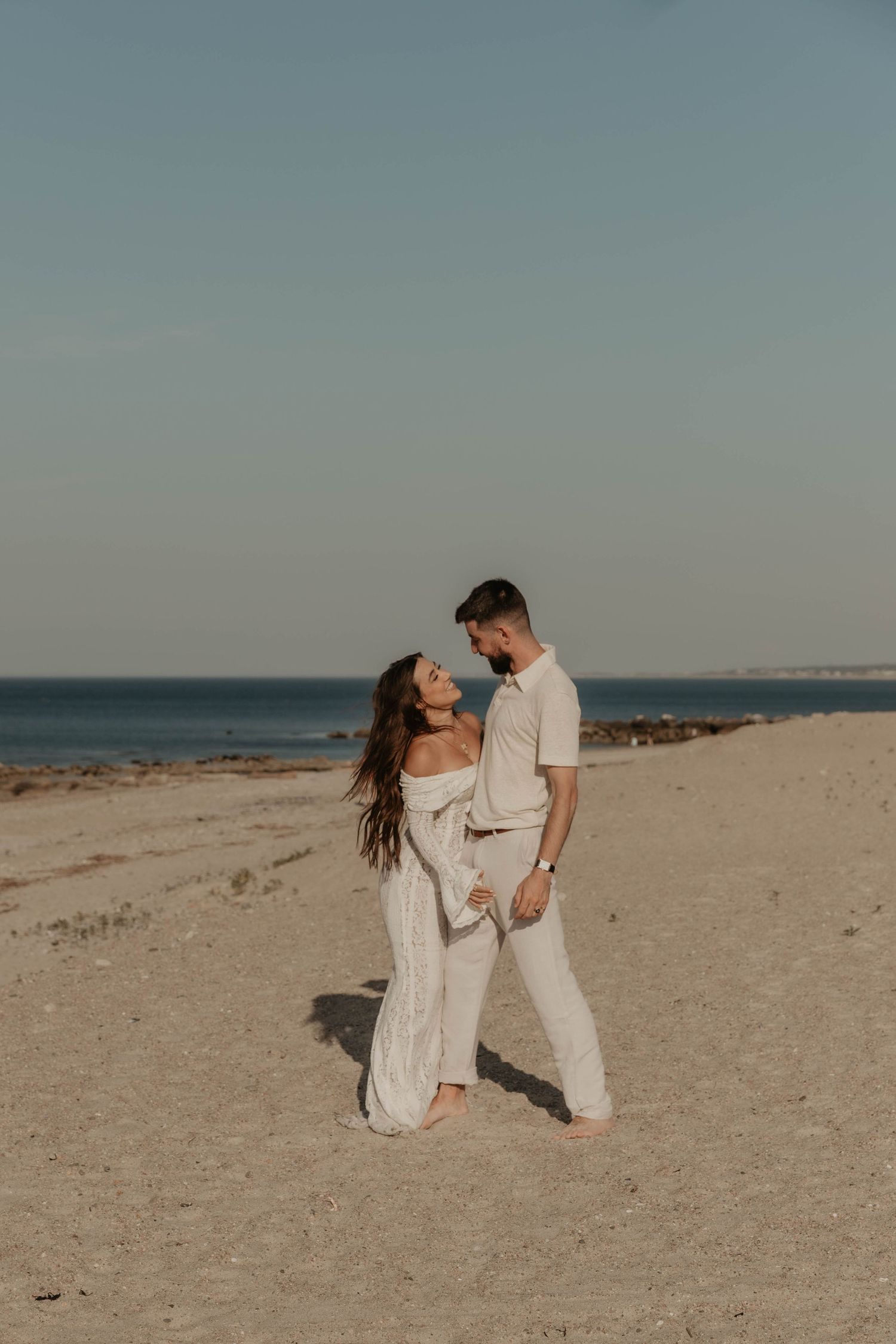Couple in matching white outfits standing barefoot on sandy beach, sharing a romantic moment with ocean in background.