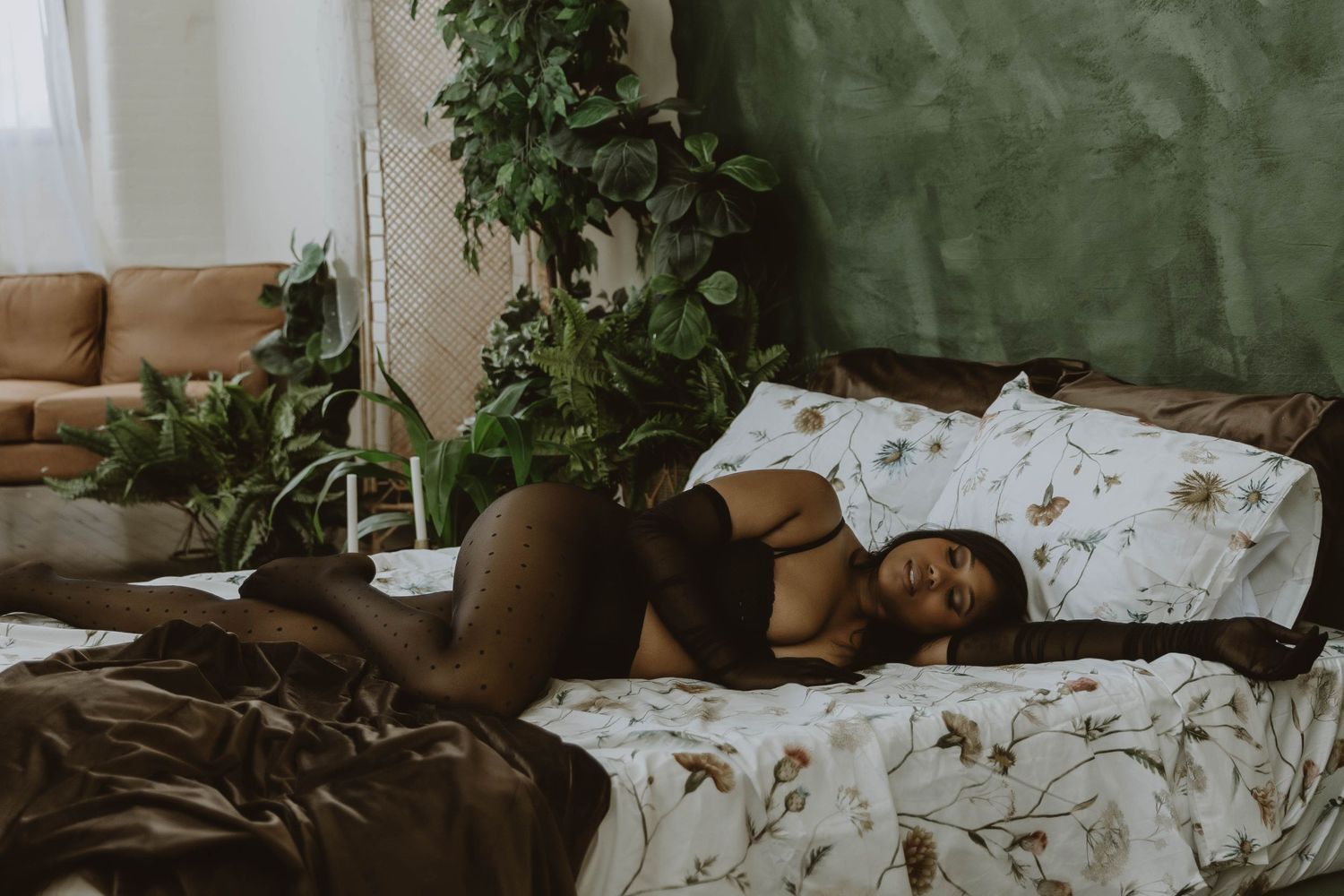 A woman in dark lingerie rests on a floral bed surrounded by lush green plants in a cozy room.