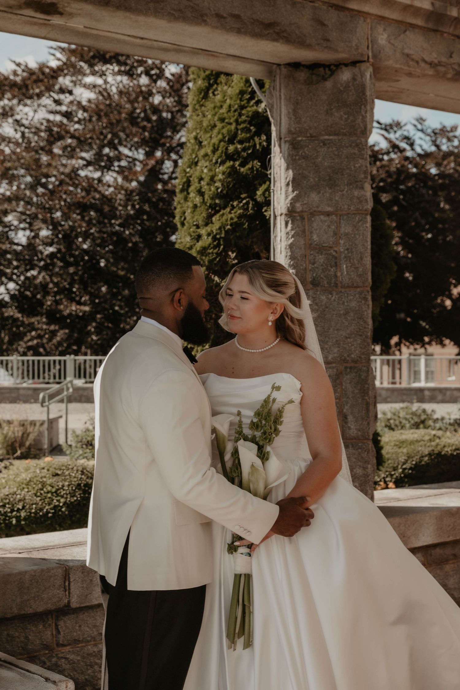 A bride and groom share a romantic moment under a stone pergola, with the bride holding a white calla lily bouquet.