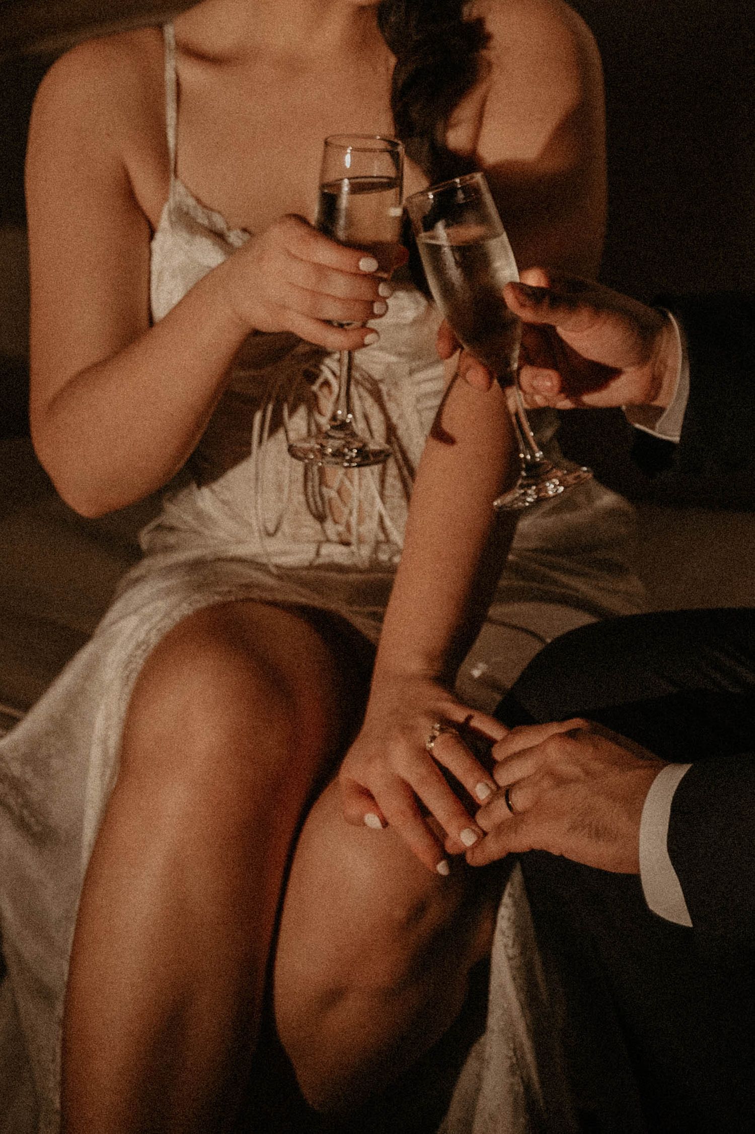Couple in warm, intimate lighting toasting with champagne flutes, her in a white satin slip dress, his hand resting on her knee.