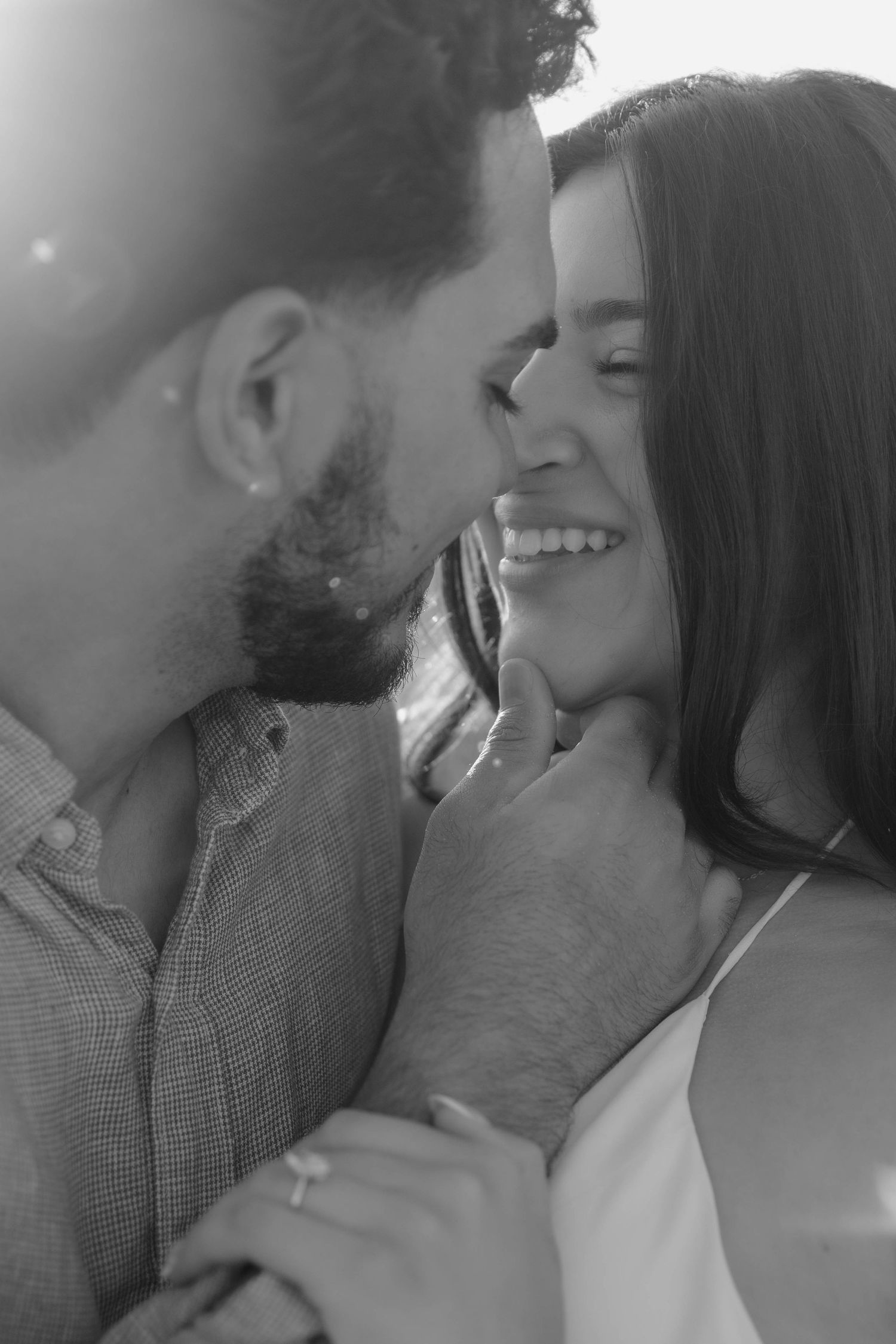 Black and white engagement photo of a smiling couple nose to nose as he gently holds her chin, her ring visible, in soft glowing light.