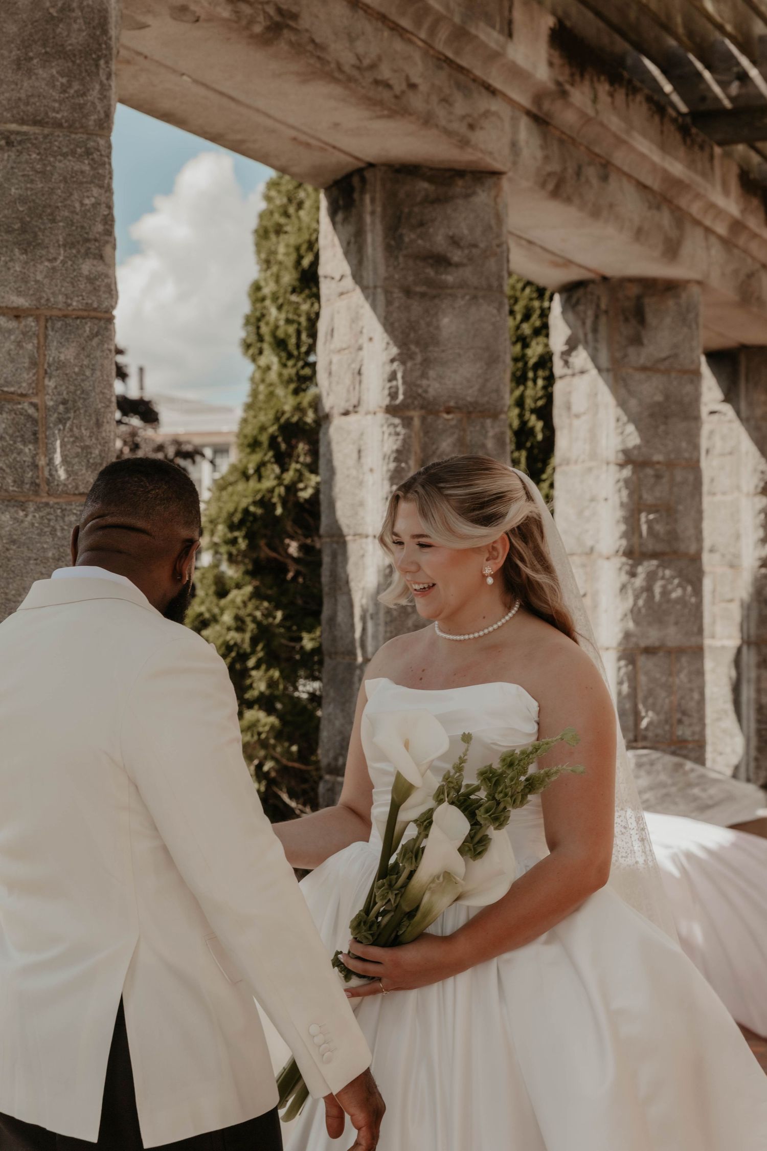 Bride in strapless white gown holds calla lily bouquet, smiling at groom in white suit near stone archway.