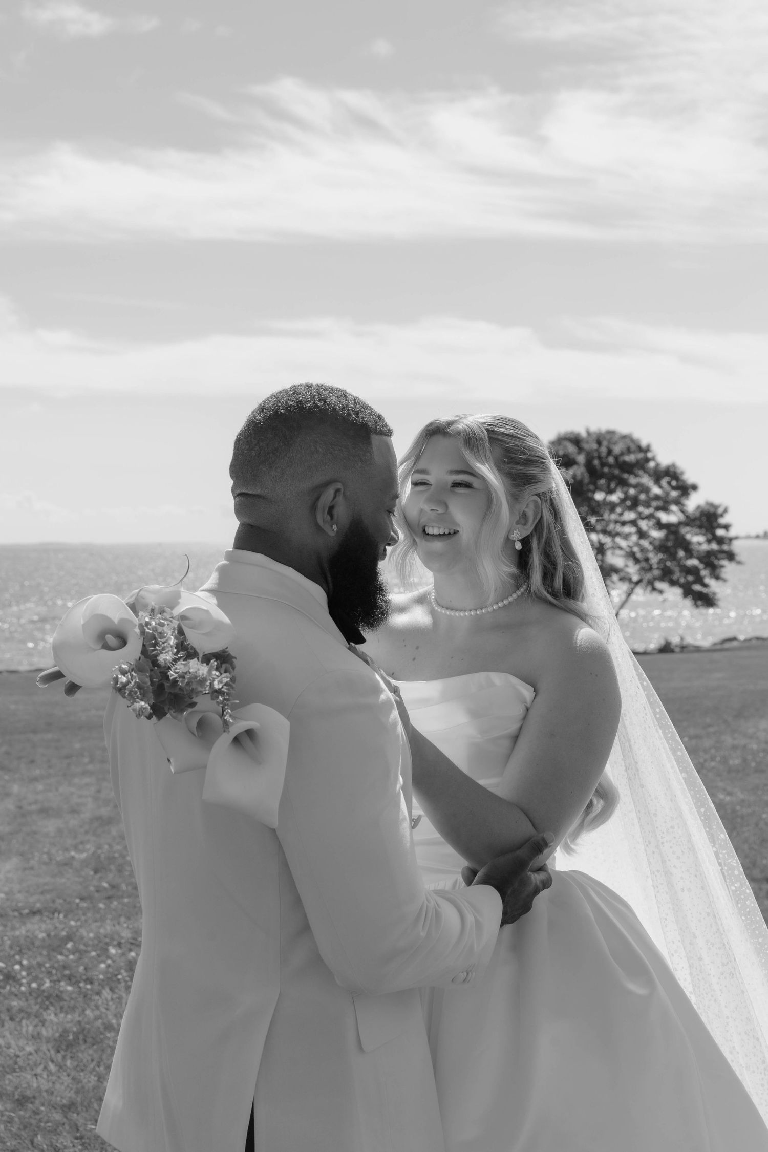 Black and white photo of a joyful bride and groom embracing outdoors on their wedding day near the ocean.