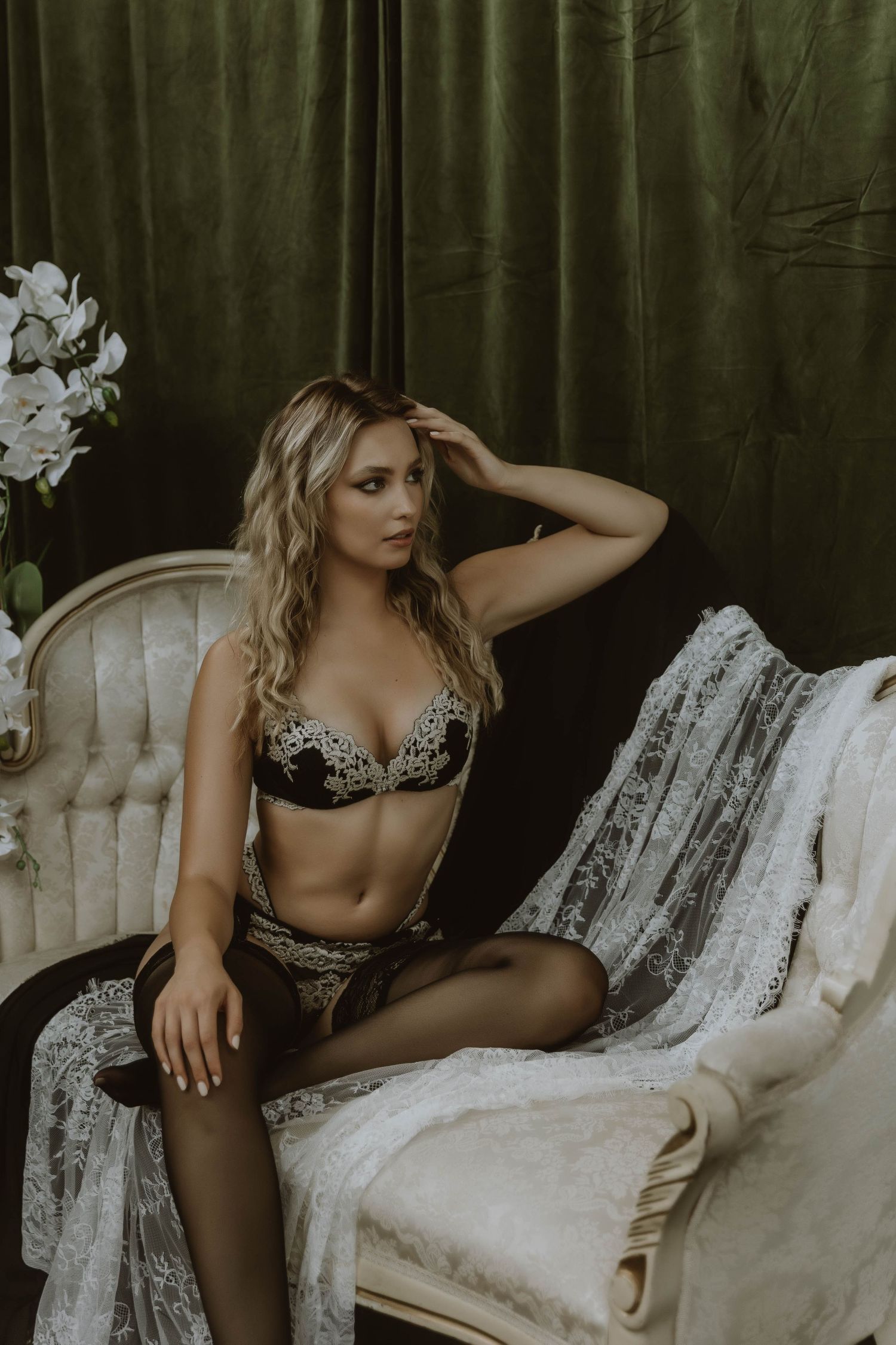 Blonde woman in gold and black lace lingerie seated on white vintage chair with green curtain backdrop.