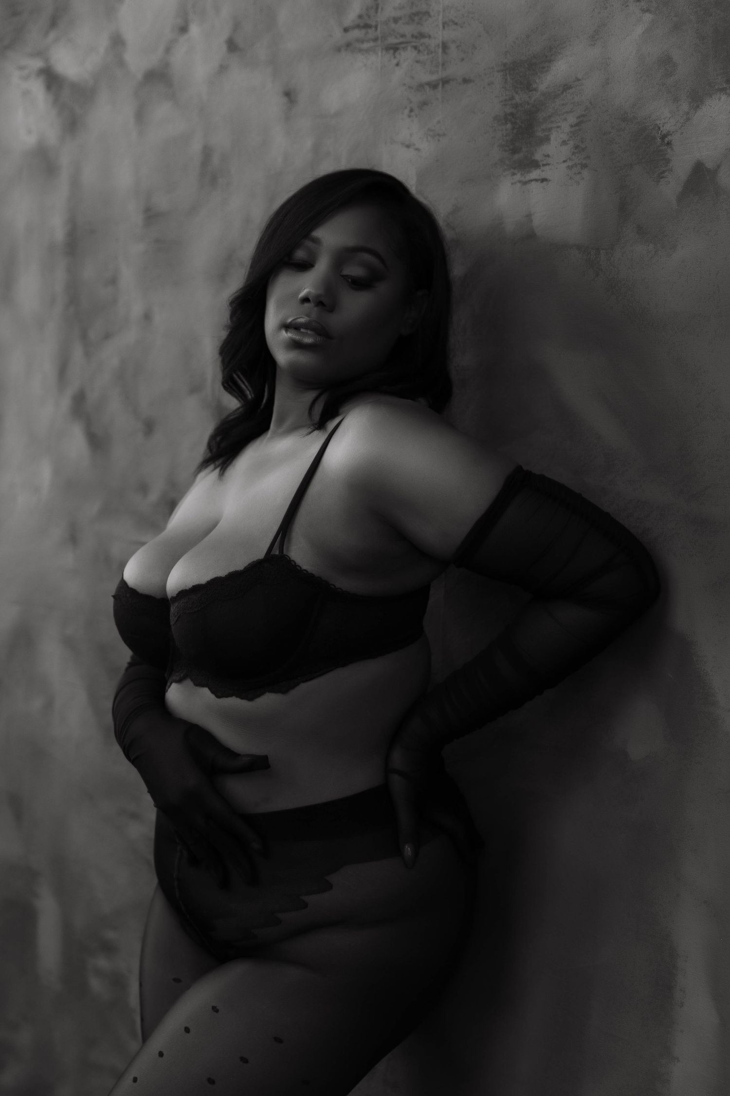 Black and white boudoir photo of a woman in lingerie and gloves leaning against a textured wall.