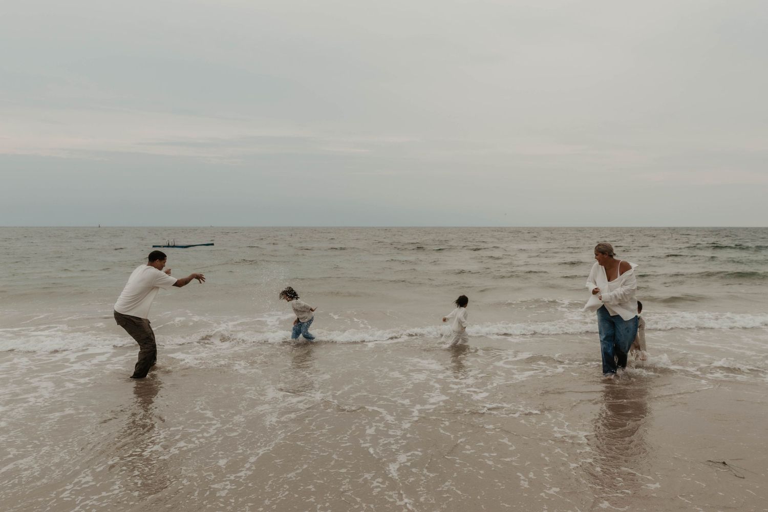 Family of four playing and splashing in shallow ocean waves on a cloudy day at a calm, wide sandy beach.