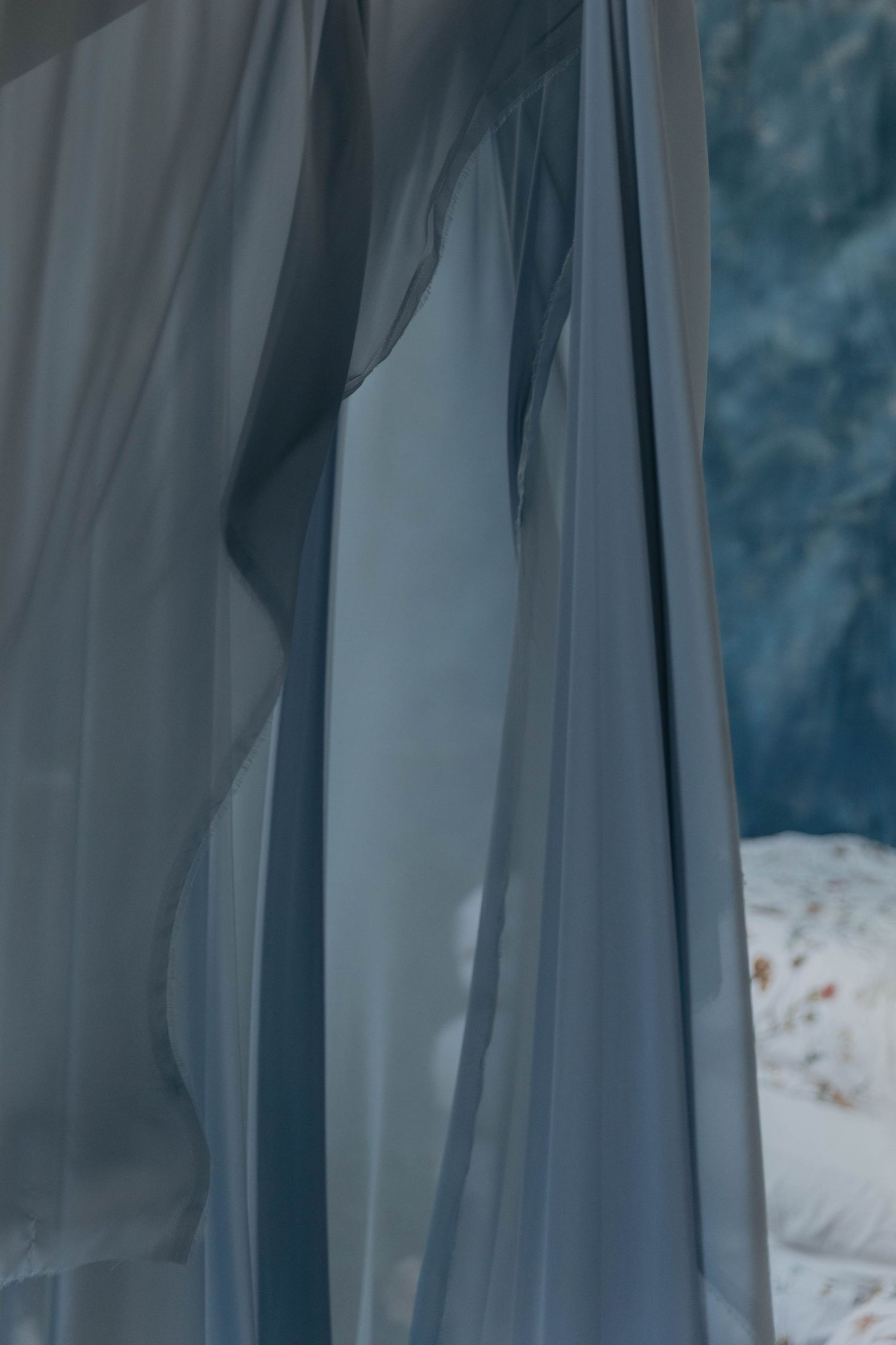 Sheer blue-gray curtains draped elegantly in a softly lit bedroom with a blurred floral bedspread in the background.