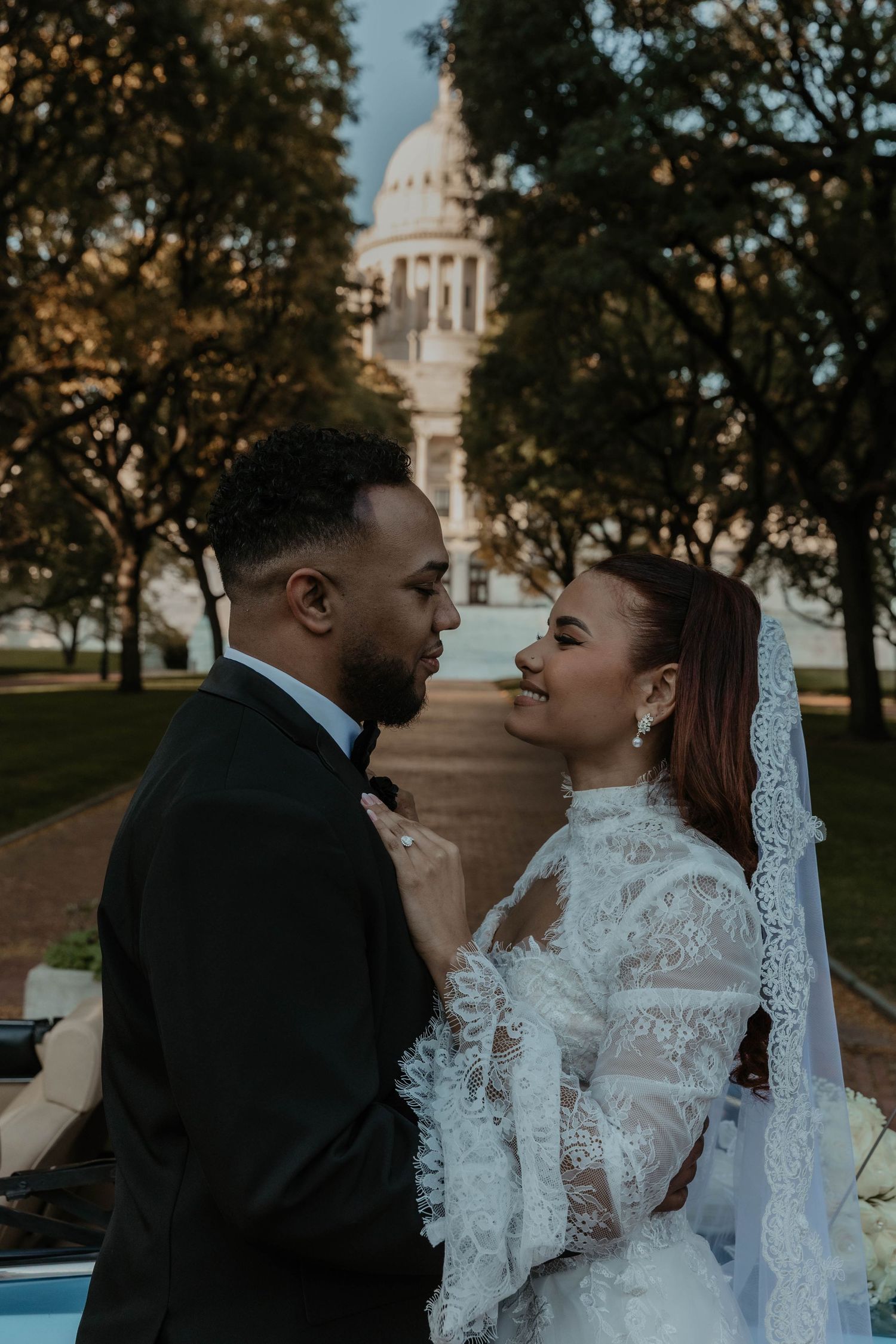 A bride in a lace gown and veil smiles at her groom in a black suit as they share an intimate moment before a domed capitol building.