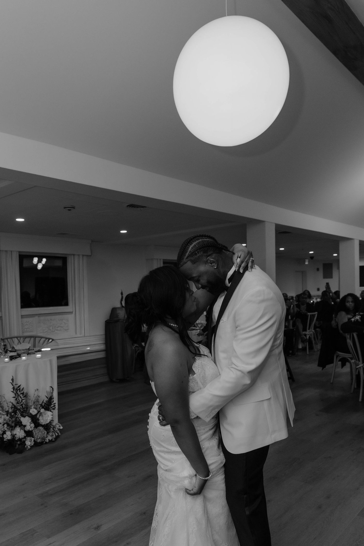 Black and white photo of a couple sharing their first dance at a wedding reception, embracing under a round pendant light.