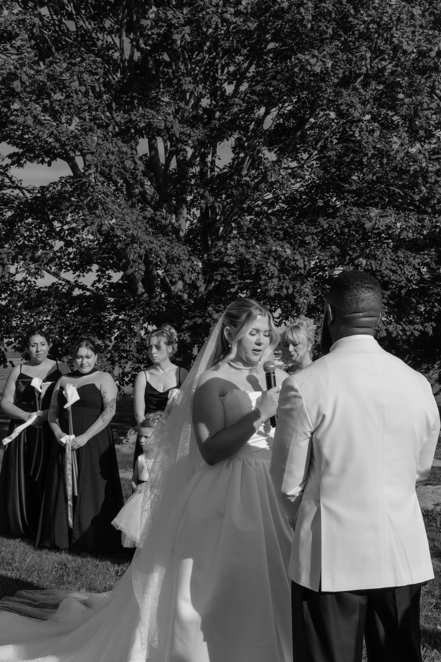 Black and white outdoor wedding ceremony with bride in gown and veil, groom in white jacket, bridesmaids in dark dresses.