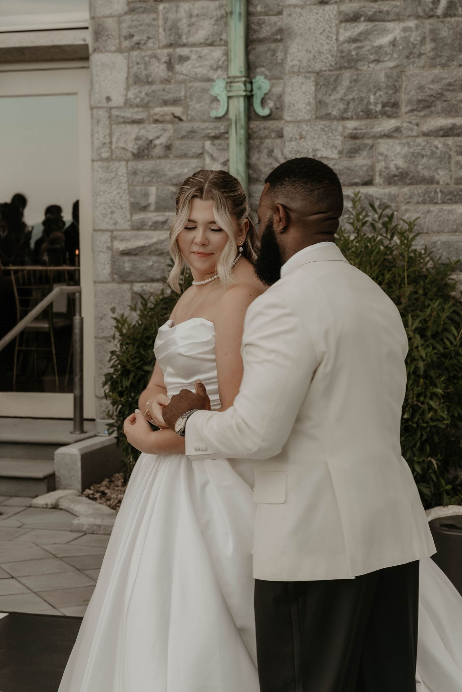 A bride in a white strapless gown and groom in a white tuxedo share a tender moment outside a stone church.