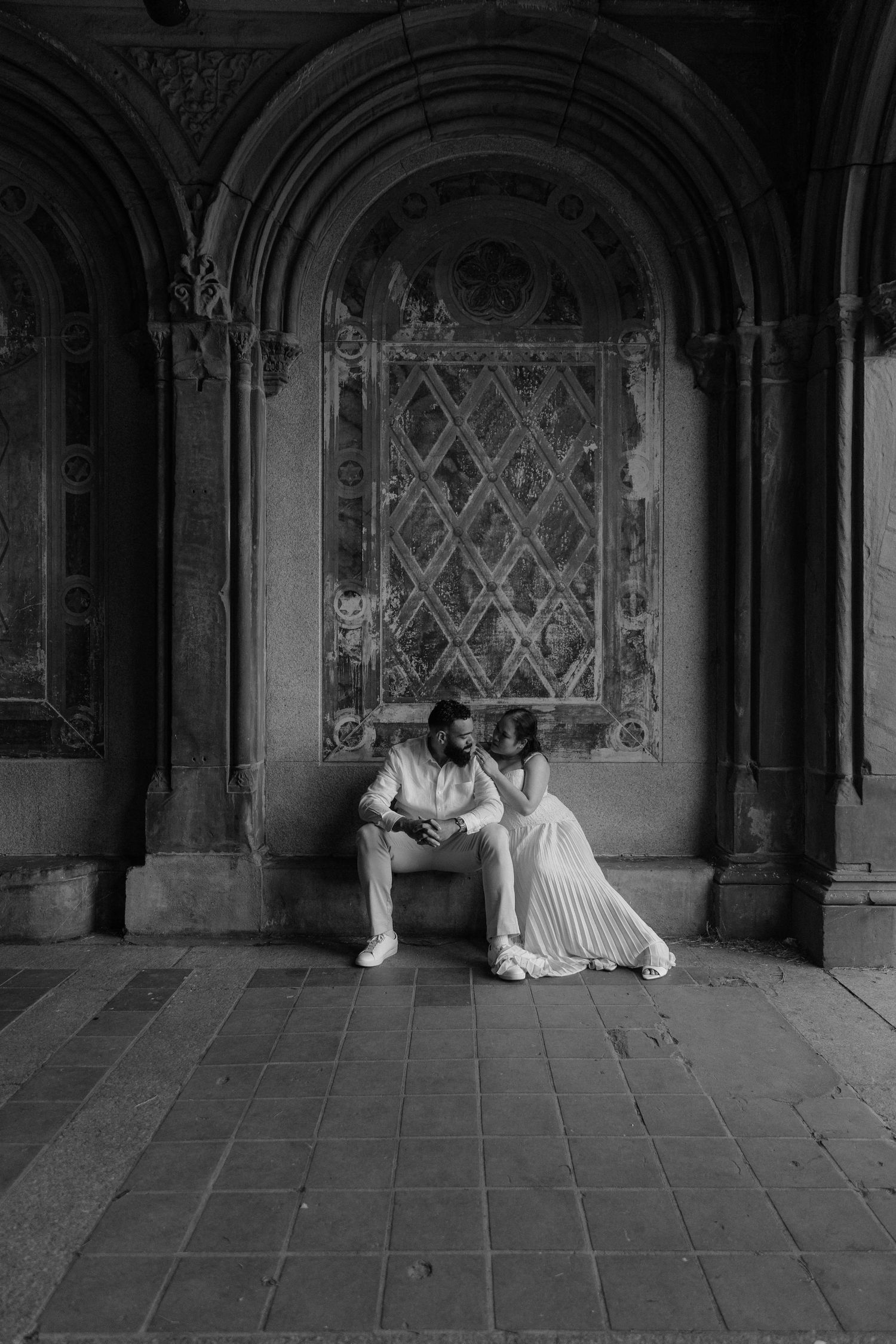 Black and white photo of a couple in wedding attire sitting together under ornate stone arches at Bethesda Terrace.