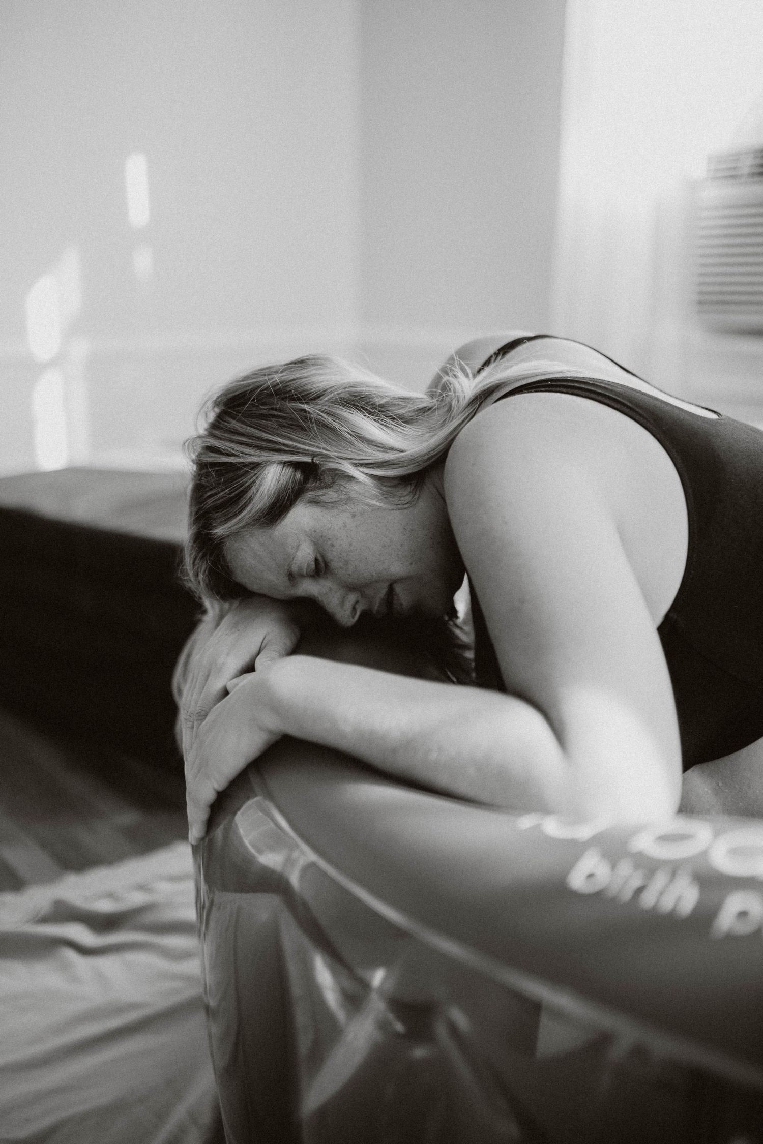 Black and white photo of a woman leaning over a birth pool during labor, resting her head on her arms.