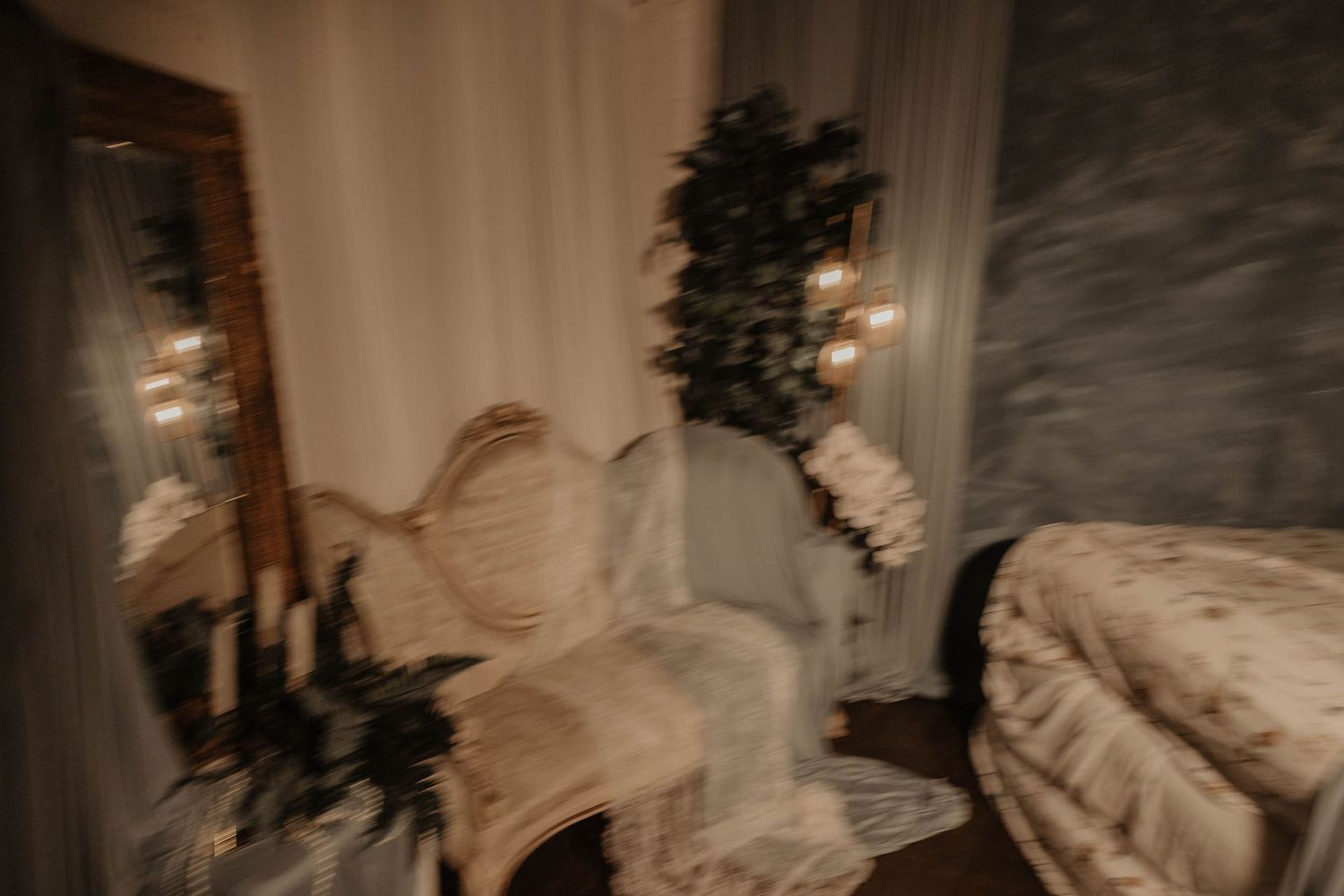 Blurry romantic bedroom vignette with ornate chair, candles, mirror, and lush greenery in warm moody lighting.