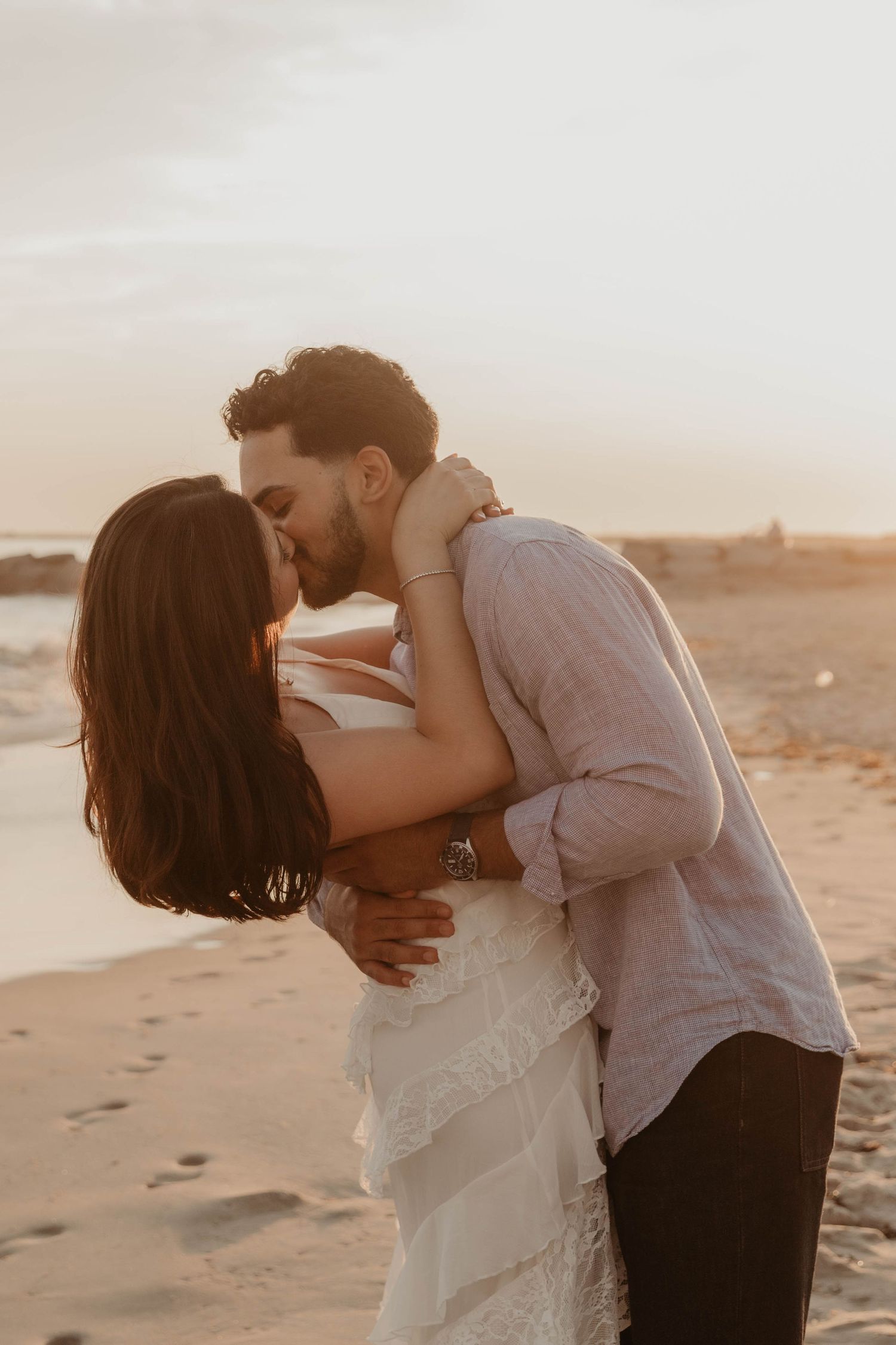 Couple sharing a romantic kiss on a sandy beach at golden sunset, woman in white dress, man in light pink shirt.