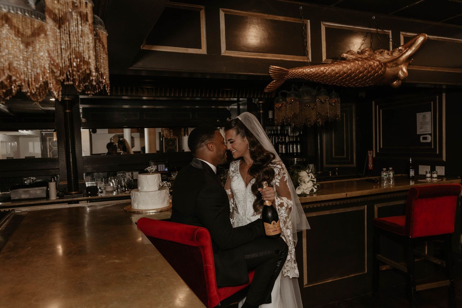 Bride and groom share romantic moment at elegant dark bar with red chairs and gold chandelier on wedding day.