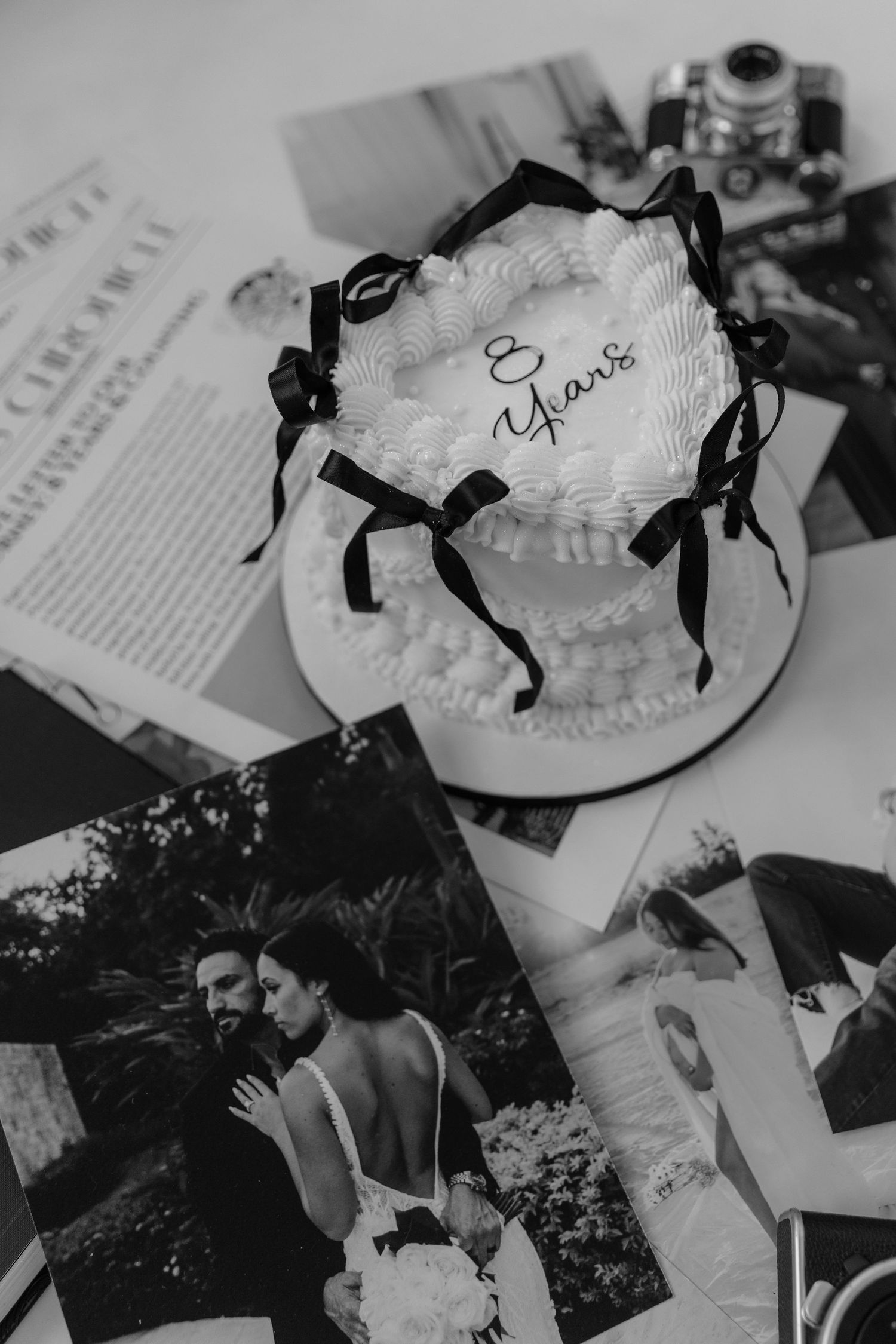 Black and white photo of an anniversary cake reading 8 Years with black bows, surrounded by wedding photos and a vintage camera.