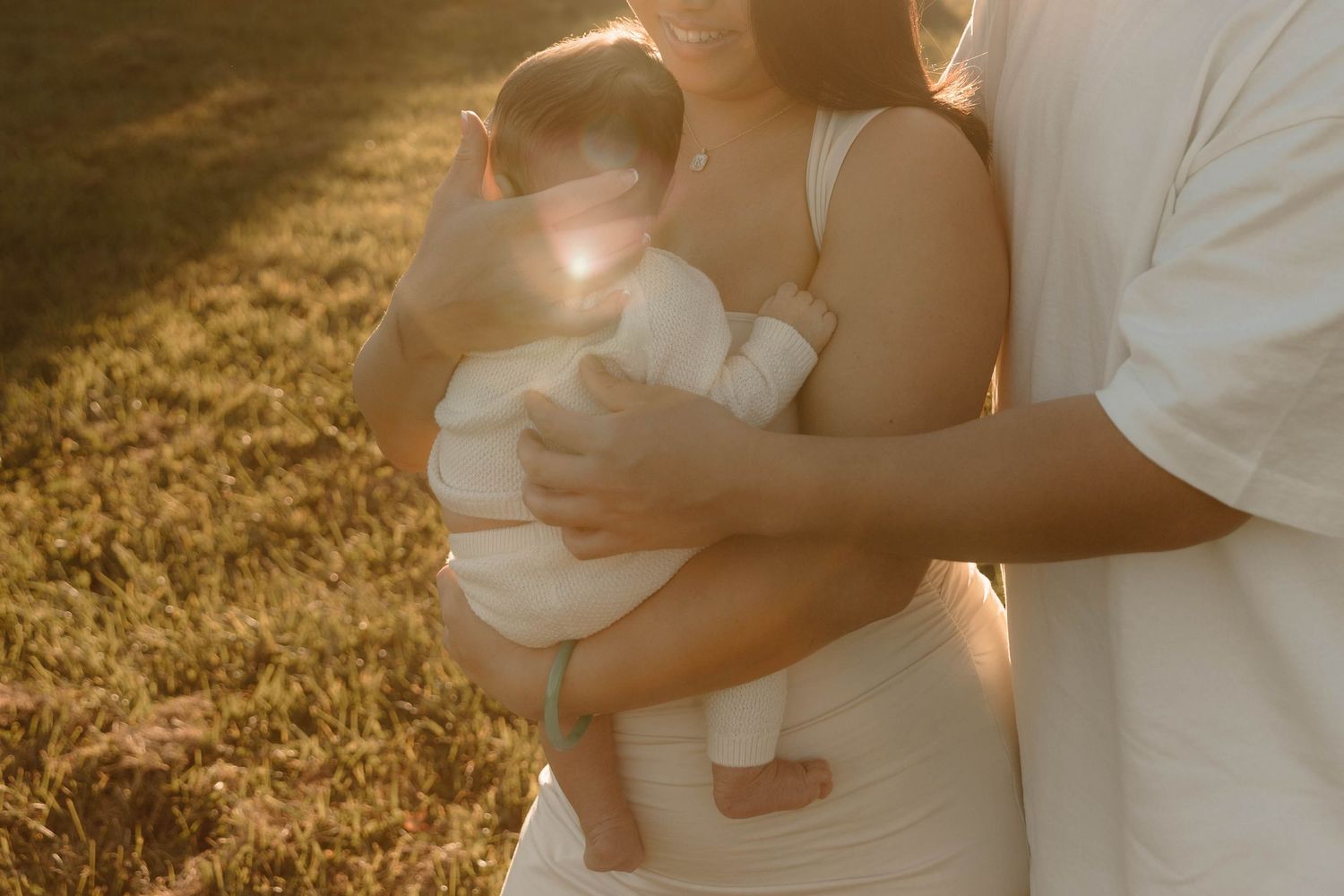 Parents in white outfits holding their newborn baby in golden sunset light outdoors.