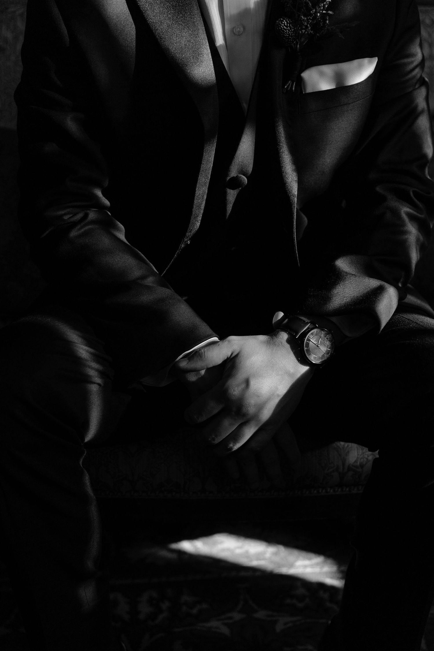 Black and white photo of a man in a suit with a watch on his wrist, sitting with hands clasped together.