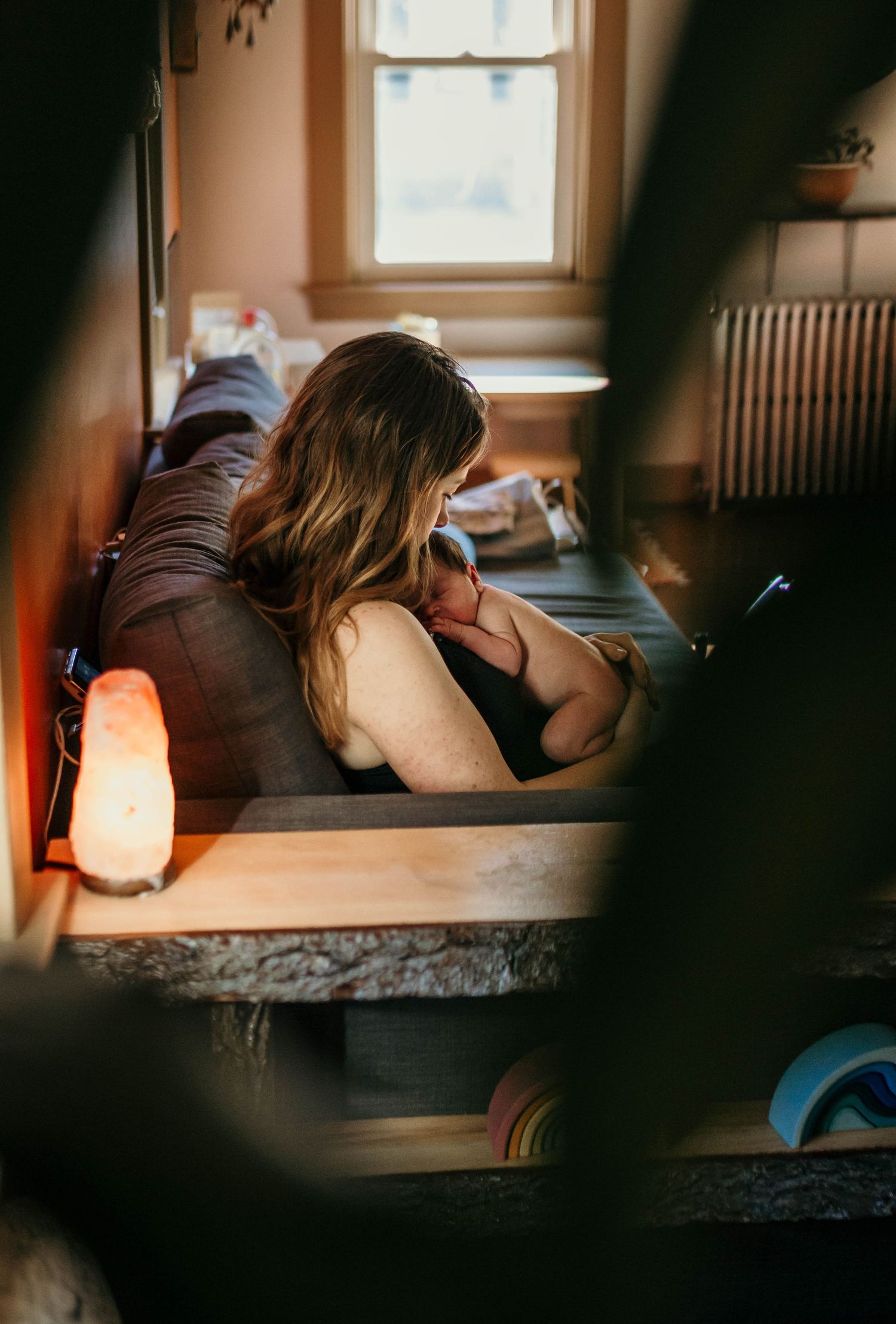 A woman cradles a newborn baby while resting on a couch near a window with warm lamp light.