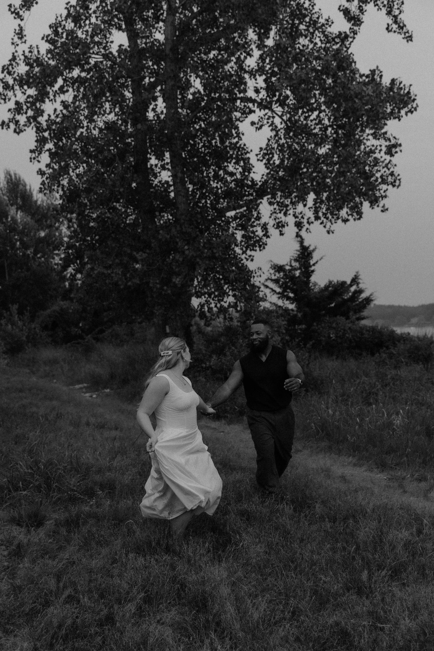 Black and white photo of a bride and groom holding hands running through a grassy field with trees in the background.