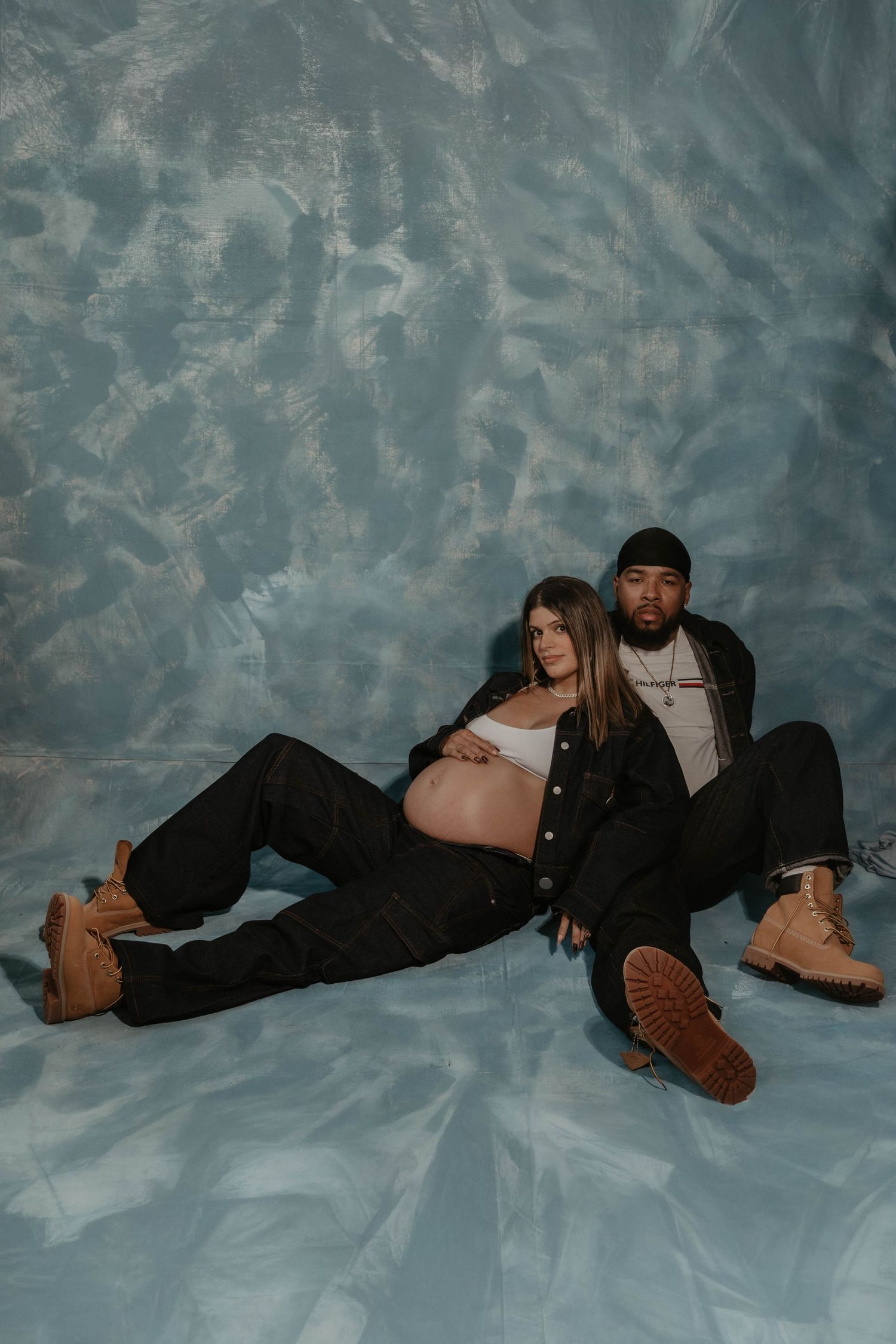 Pregnant couple posing on blue backdrop, woman in white crop top and black pants, man in dark jacket and tan boots.