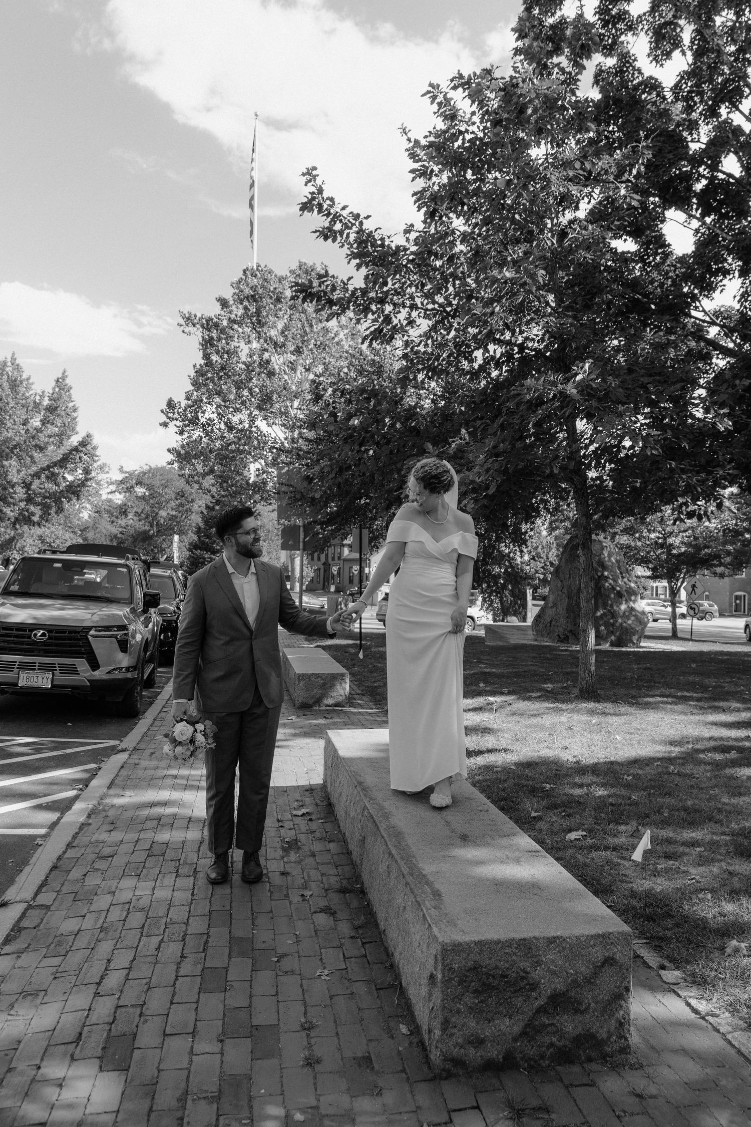 Black and white photo of a bride and groom walking together outdoors on a brick path near a park on their wedding day.