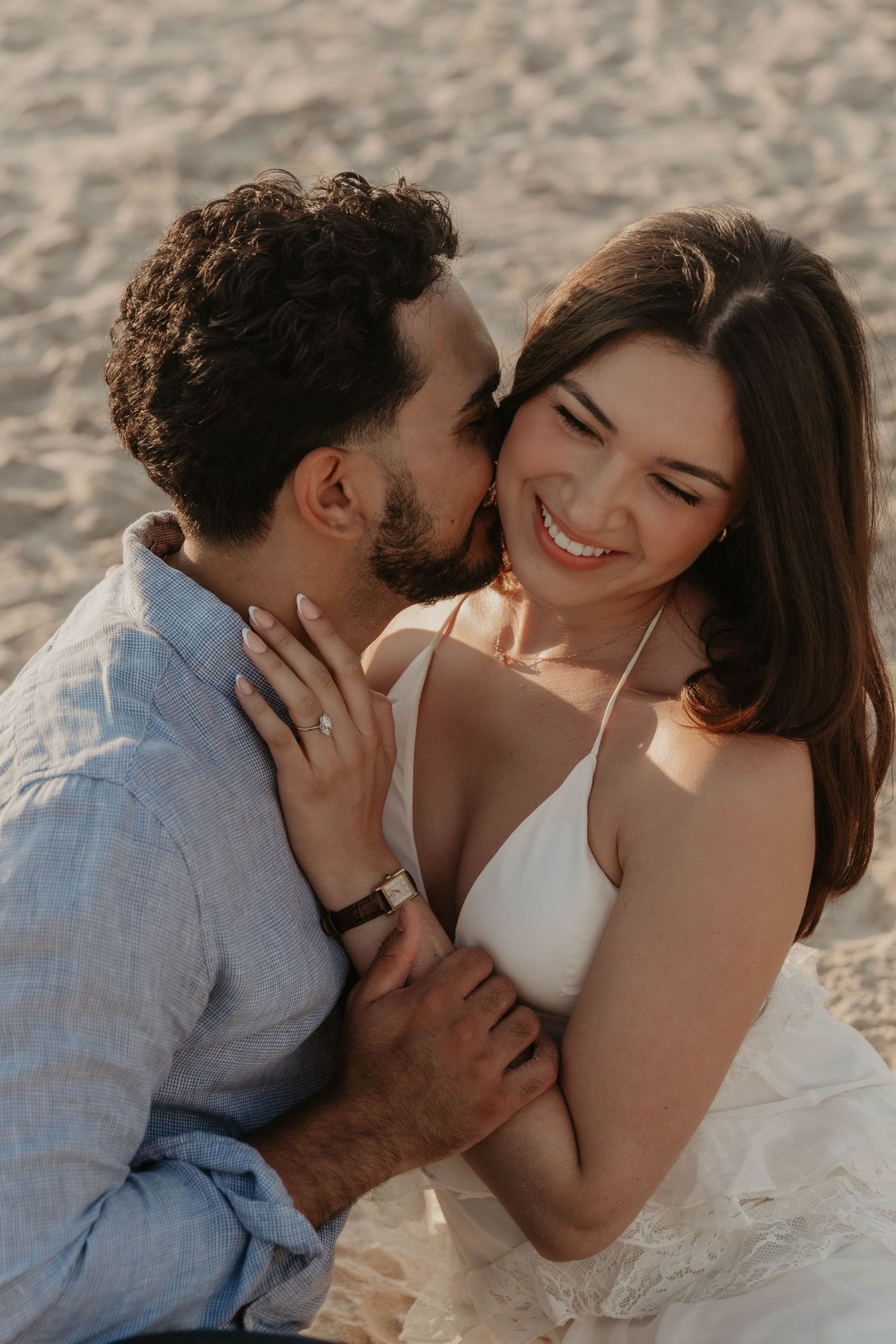 A couple shares a romantic beach moment, man kissing woman's cheek as she smiles joyfully, showcasing an engagement ring.