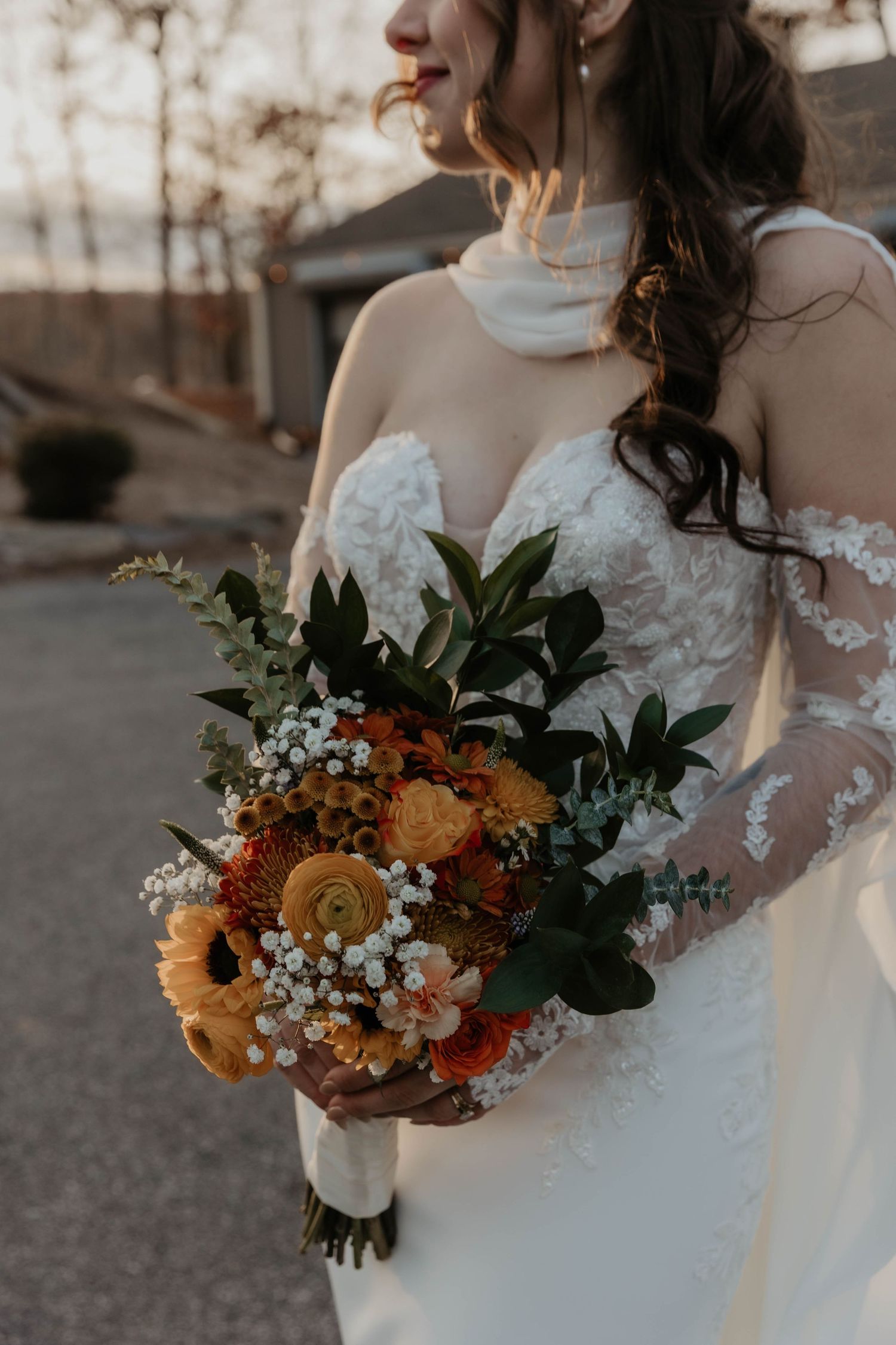 Bride in a white lace off-shoulder gown holding an autumn bouquet of orange ranunculus, white baby's breath, and greenery outdoors.