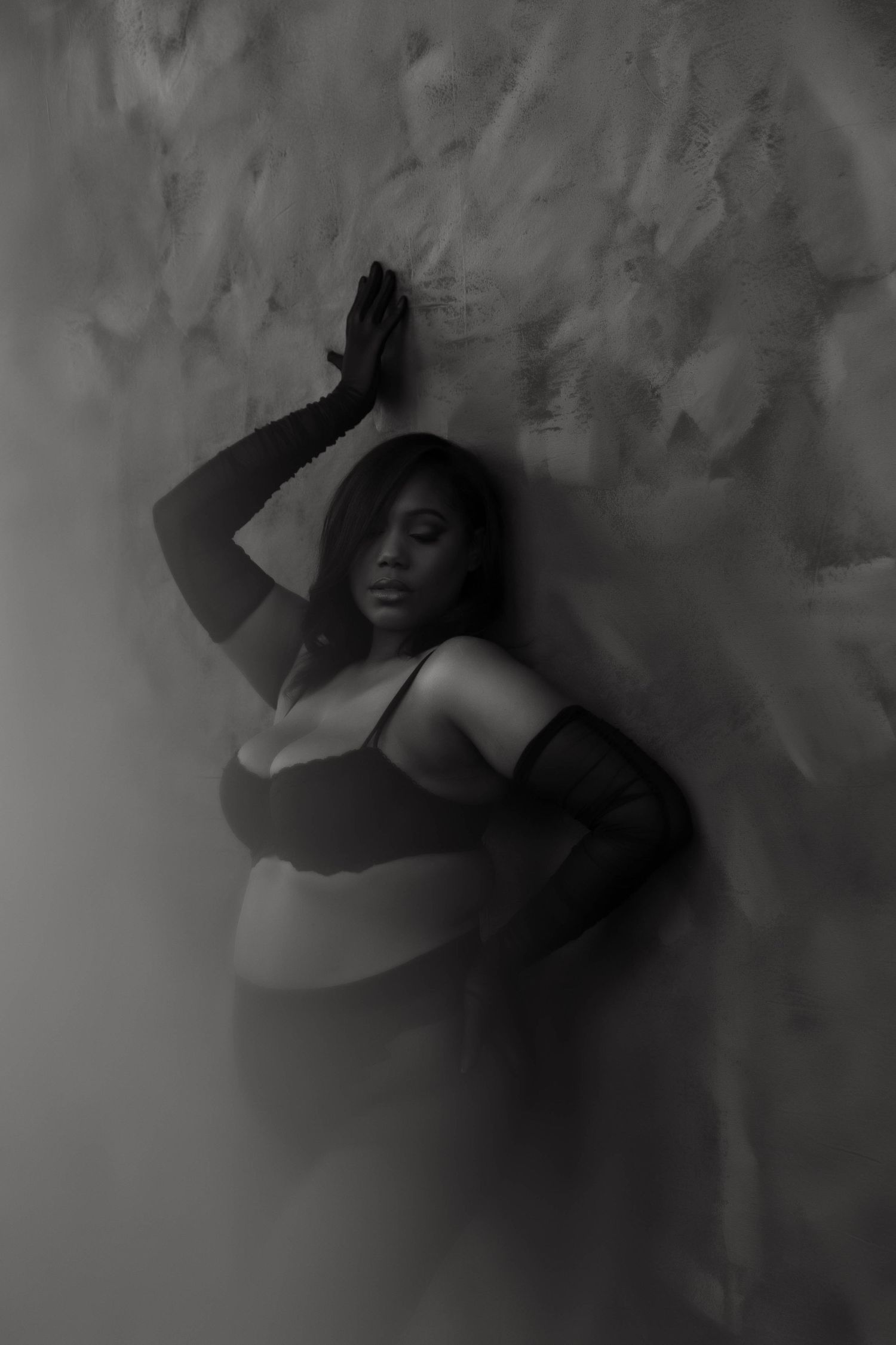 Black and white boudoir photo of a plus-size woman in lingerie leaning against a textured wall in dramatic lighting.