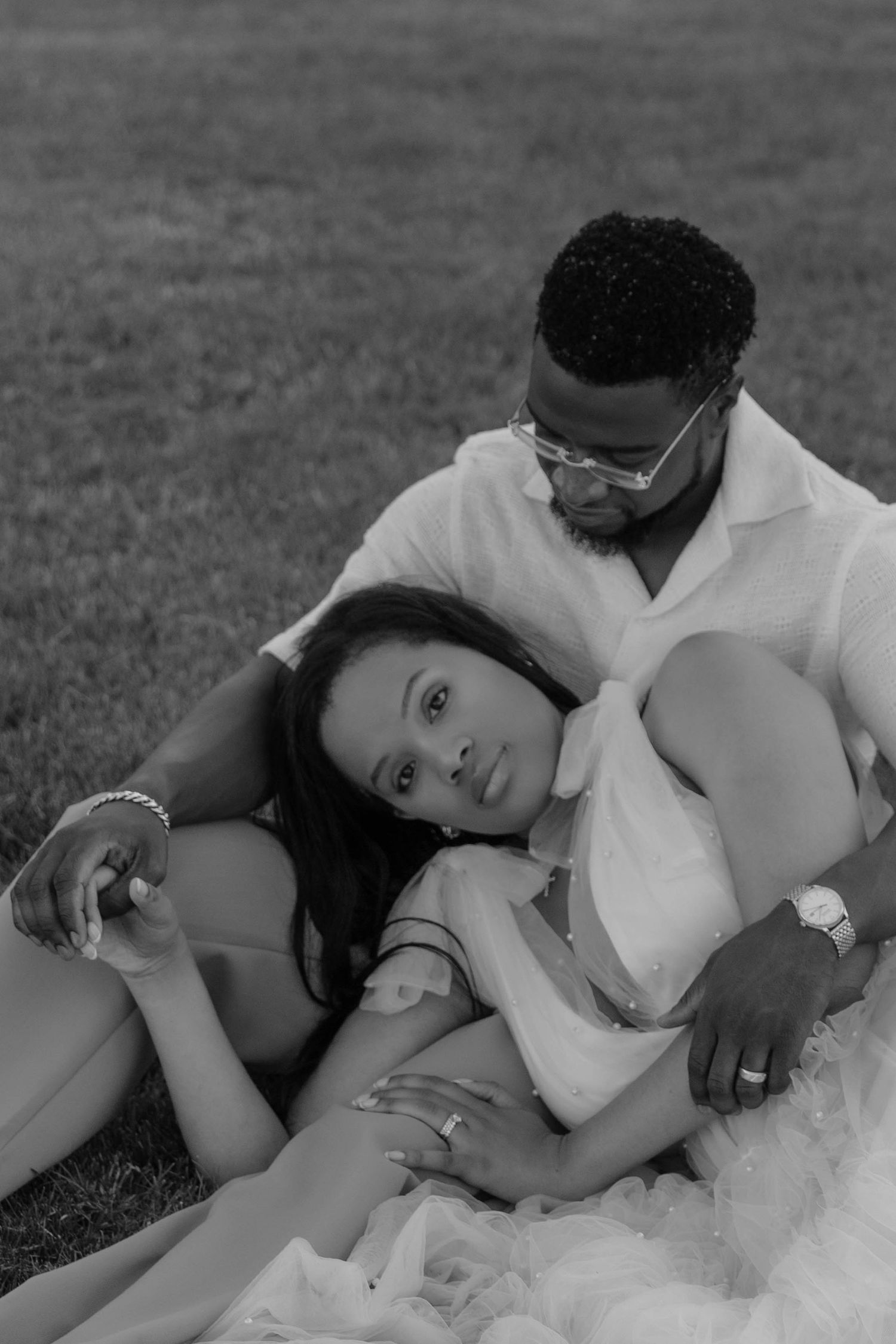 Black and white photo of a couple relaxing on grass, woman leaning on mans chest in a tender embrace.