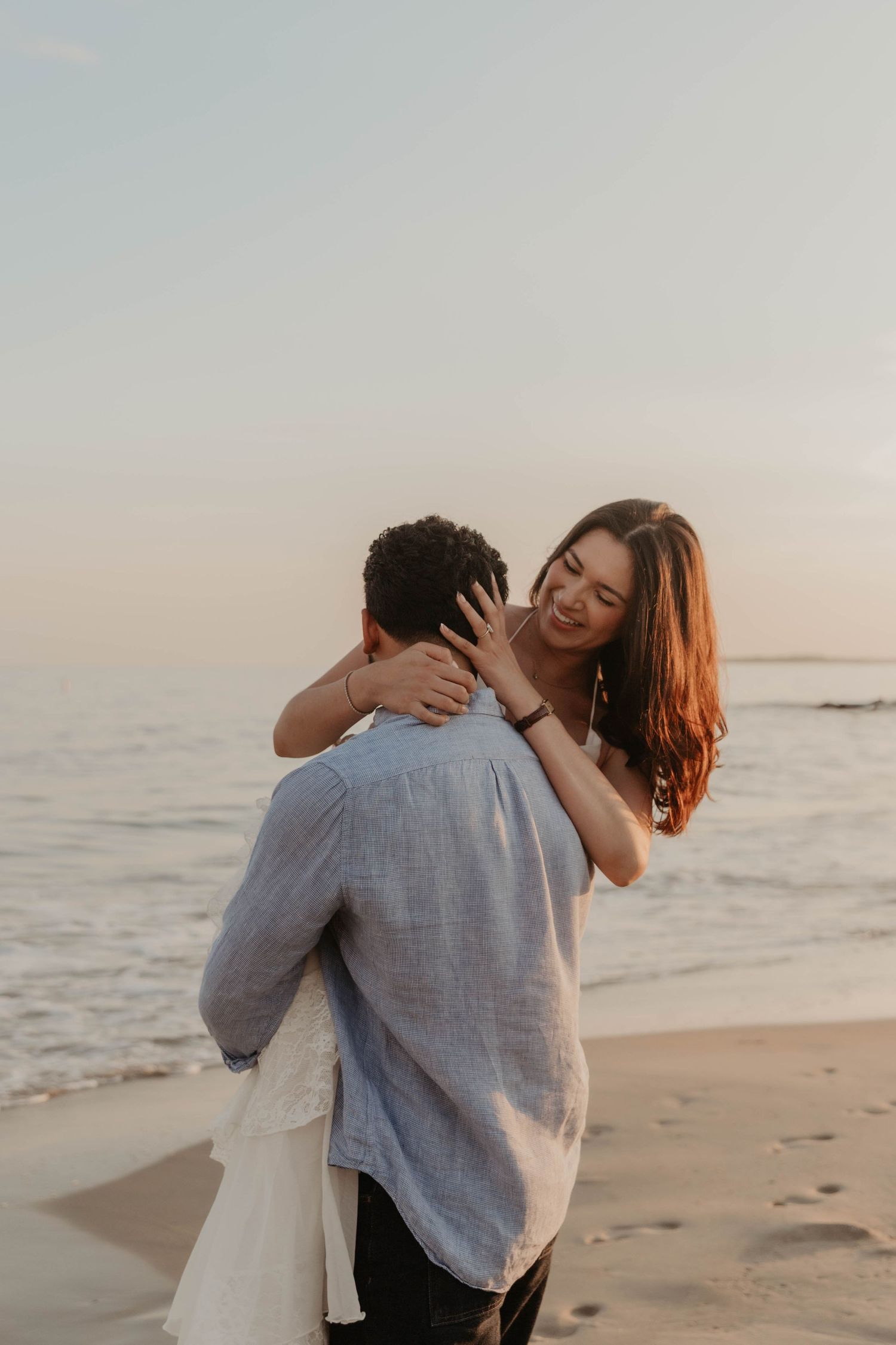 Couple embracing romantically on a beach at sunset, woman smiling with arms around man's neck by the ocean.
