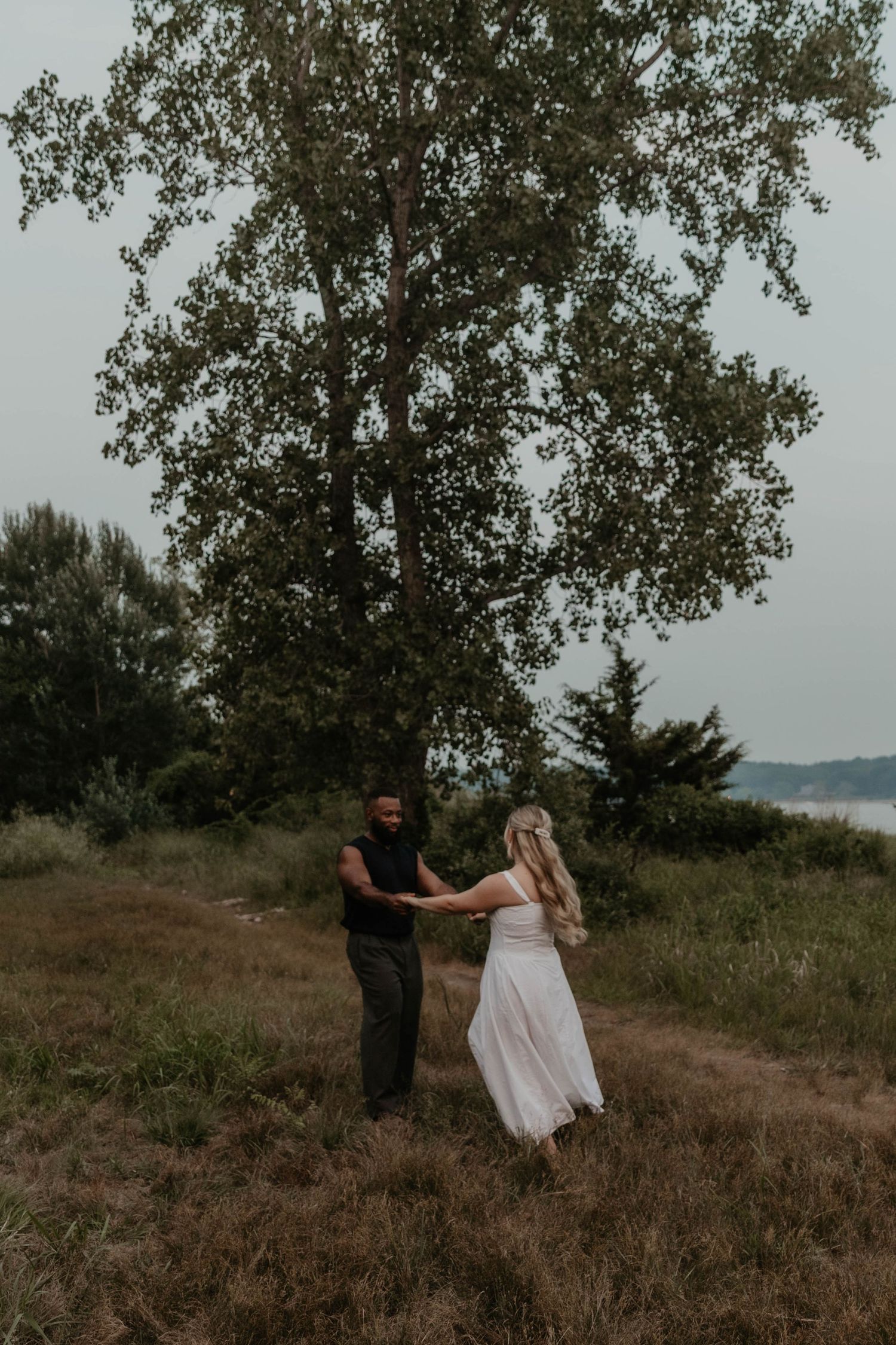 Couple holding hands in a field near a large tree, woman in white dress and man in black, romantic outdoor photo.